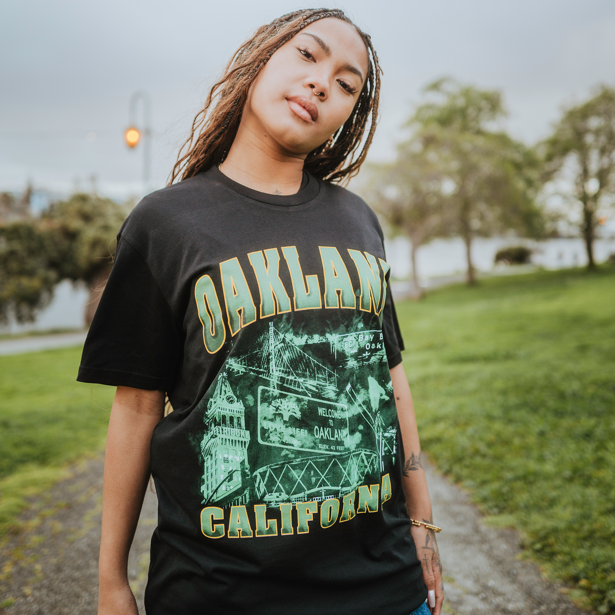A woman with braided hair confidently wears a classic fit Montage Tee by Oaklandish in black, featuring intricate city imagery. She stands on a grassy path, trees blurred behind her, embodying relaxed Emerald City vibes with her hands casually on her hips.