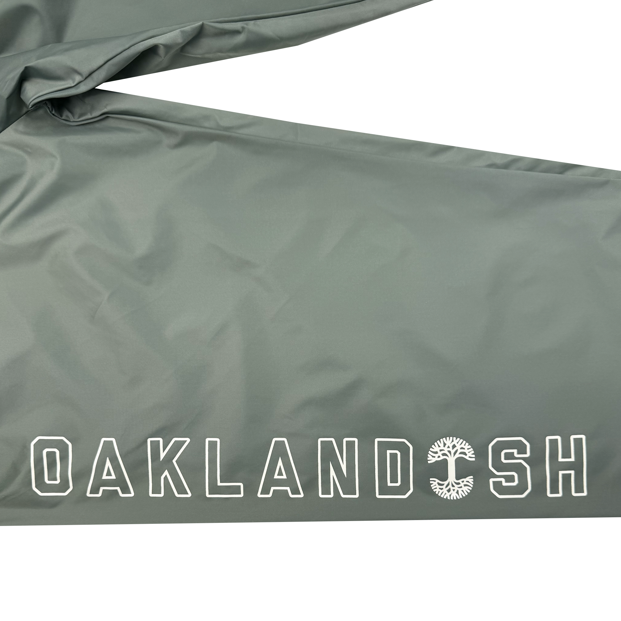 A gray fabric with "OAKLANDISH" in white capitals graces the bottom right corner, and the letter "D" is adorned with a stylized tree design. The Bandit Nylon Pant, designed by Oaklandish, features an adjustable waistband and baggy fit, giving it a slightly wrinkled appearance at the top.