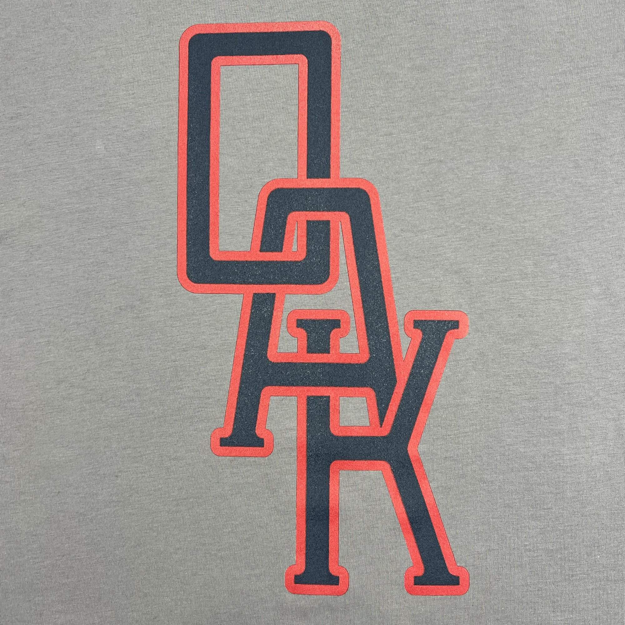 Detailed view of a grey t-shirt with OAK monogram design centered in Navy and red.
