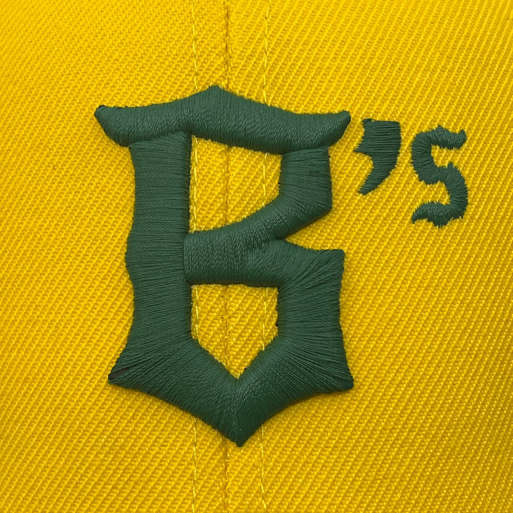 A close-up image of the Oakland Ballers Snapback showcases an embroidered logo on yellow fabric. The logo features a stylized, capital letter "B" in green thread, with an embroidered green apostrophe and letter "s" to the upper right. The yellow fabric, representing the Oakland Ballers brand, has a woven texture.