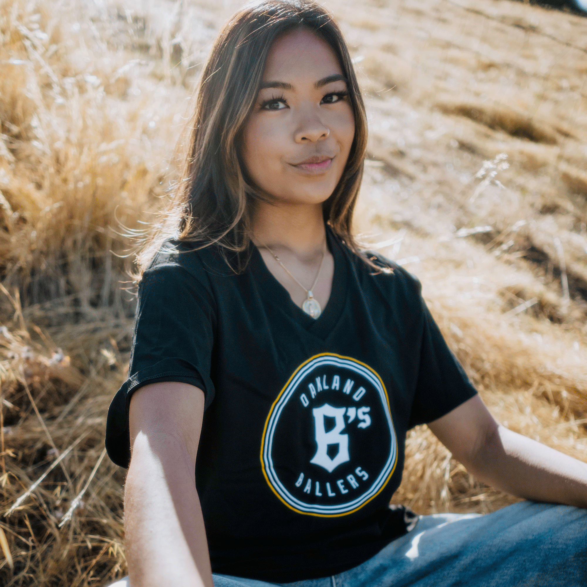 A young woman with long brown hair, wearing an Oakland Ballers Women's Logo Relaxed V Neck Tee, sits outdoors on dry, straw-like grass. She has a serene expression as the sunlight illuminates her face and surroundings.