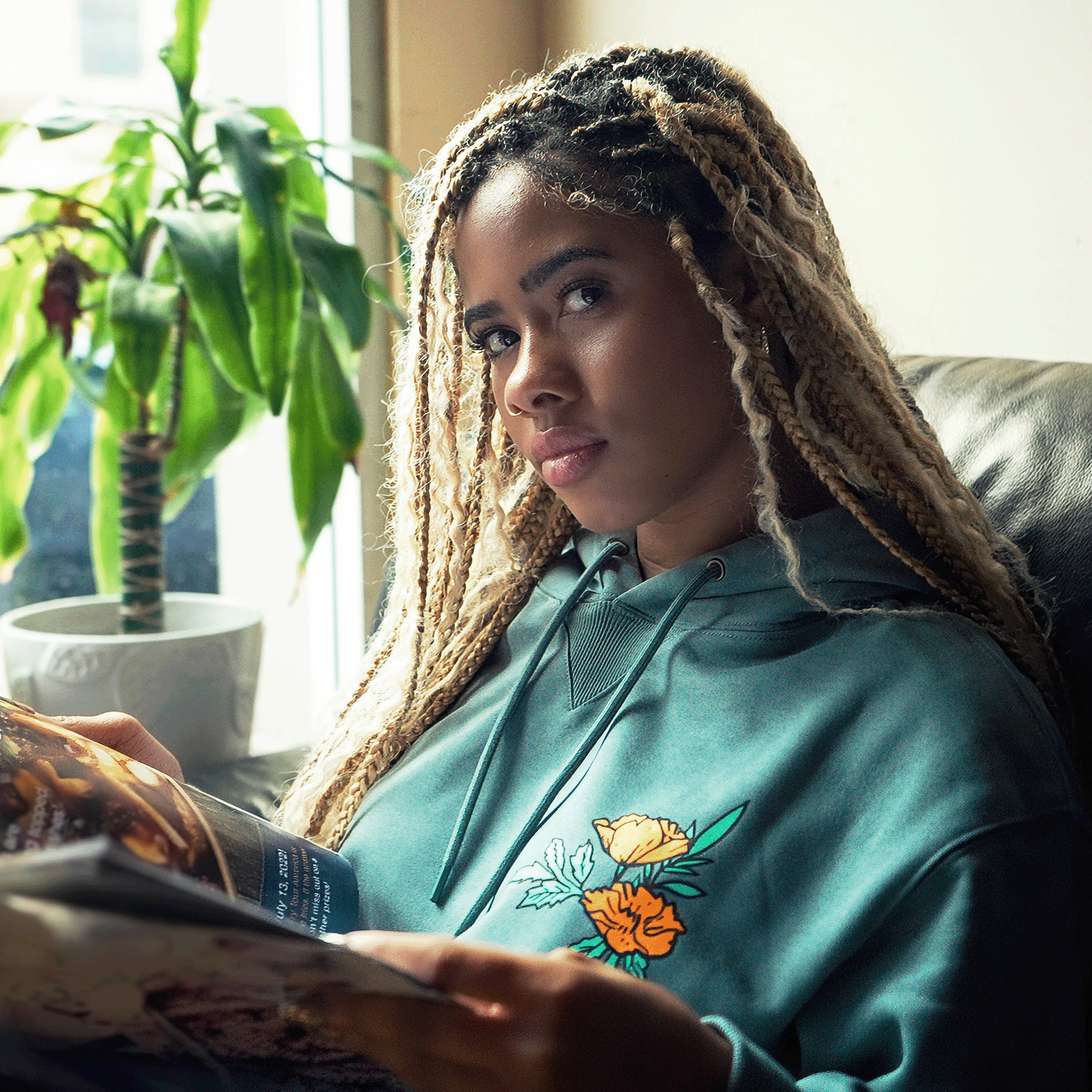 A woman with long, blonde braided hair sits on a couch near a window, holding an open magazine. She is wearing the Oakland Dream Premium Hoodie by Oaklandish, a green pullover with a floral design made of luxe French terry. Behind her is a potted plant with broad leaves. She gazes at the camera with a calm expression, illuminated by natural light.