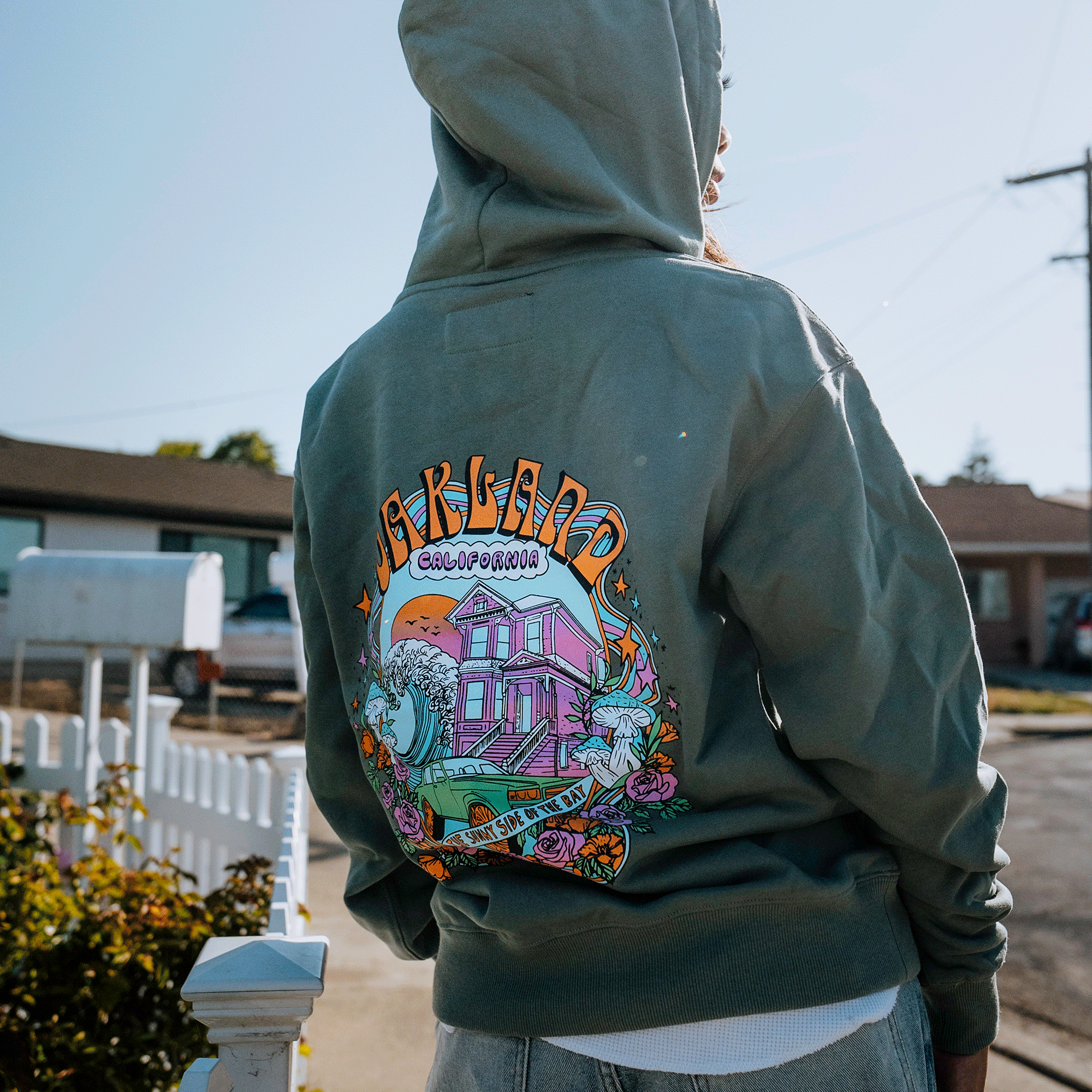 A person with long hair is standing outside wearing a green Oakland Dream Premium Hoodie from Oaklandish. The back of the hoodie features a colorful graphic reading "Oakland, California," depicting a pink house with the text "stay for the sunset." Behind them are suburban houses, a white mailbox, and a white picket fence.