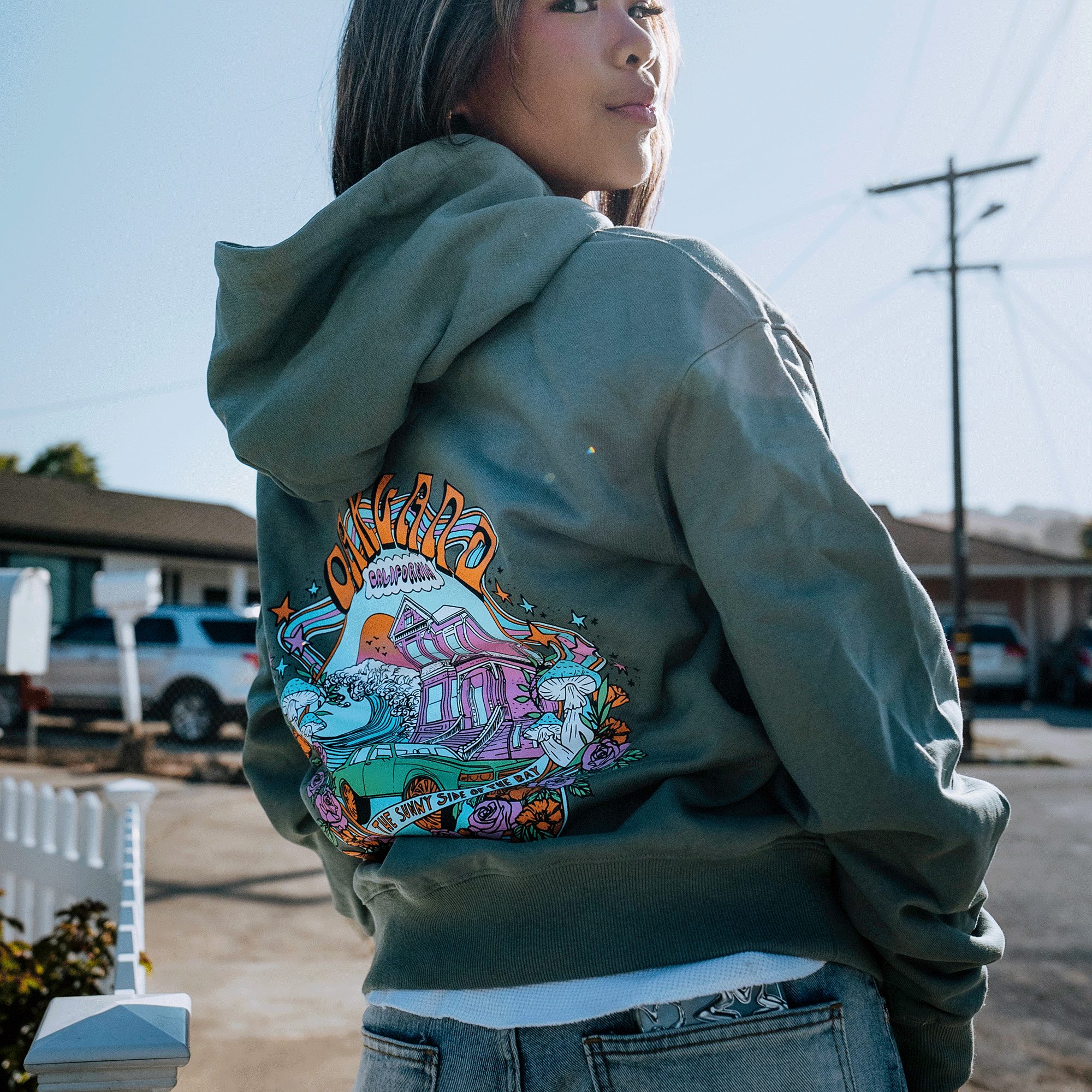 A woman with long hair stands outdoors, facing slightly away from the camera. She is wearing a green Oaklandish Oakland Dream Premium Hoodie adorned with a colorful illustration on the back, depicting a beach scene with waves, palm trees, and a setting sun, along with the word "Oxnard." The background includes houses and utility poles.