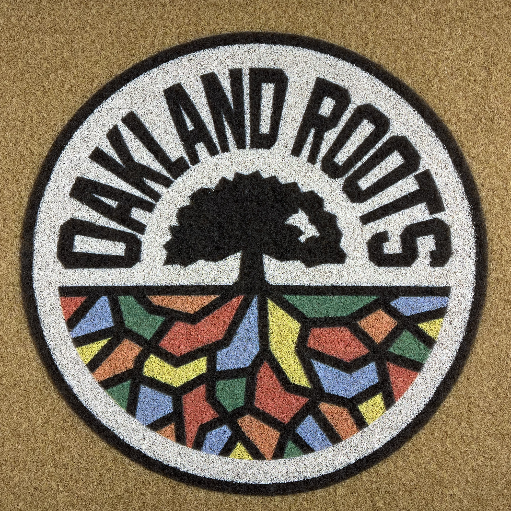 The image shows the logo of the Oakland Roots SC, as featured on the Oakland Roots SC Door Mat. The logo depicts a black tree with visible roots inside a circle. Arched over the top of the circle is the text "OAKLAND ROOTS." The lower half of the circle is filled with vibrant geometric shapes, reminiscent of stained glass, symbolizing social good.