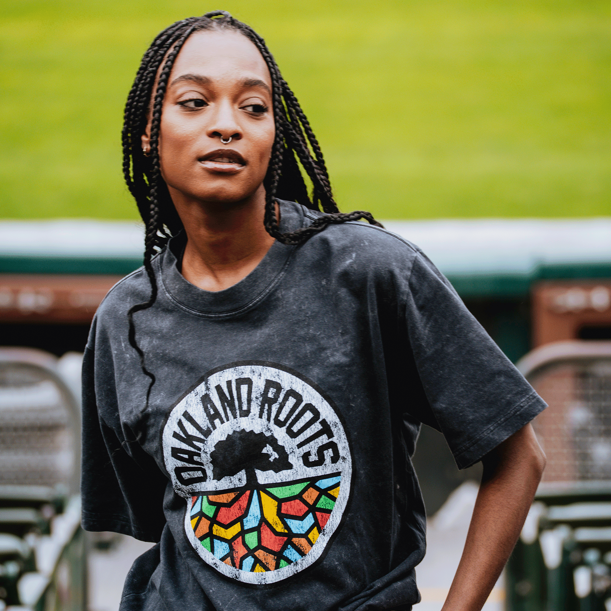A woman with long braids and a nose ring wears the Oakland Roots SC Classic Heavy Stone Wash Tee by Oakland Roots SC, featuring a tree and colorful roots. She stands outside near stadium seats, with a blurred green field behind her.