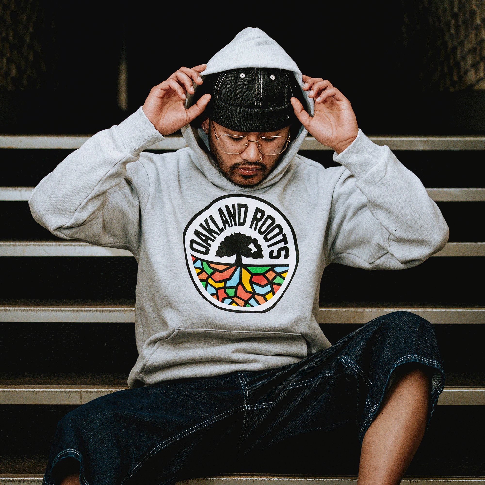 A person in glasses, black cap, and the Oakland Roots SC Heavy Hoodie by Oakland Roots SC with a colorful tree logo sits on outdoor steps, adjusting their hood and wearing dark shorts—showing local sports club pride against a neutral background.