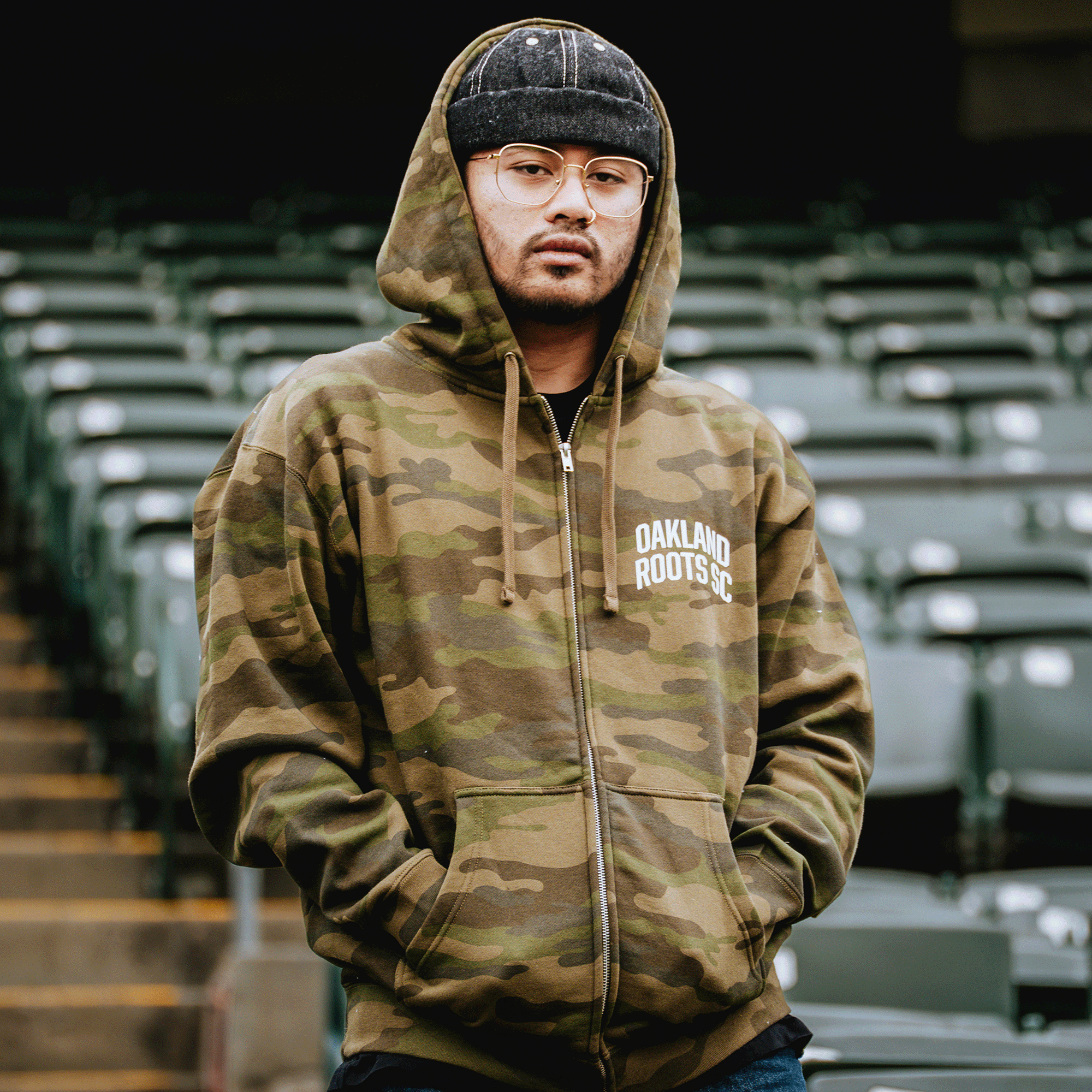 A person stands among empty green stadium seats, wearing the Oakland Roots SC Classic Zip Hoodie by Oakland Roots SC, along with a black knit beanie and glasses. Embracing classic Oakland sports style, they keep their hands in their pockets.