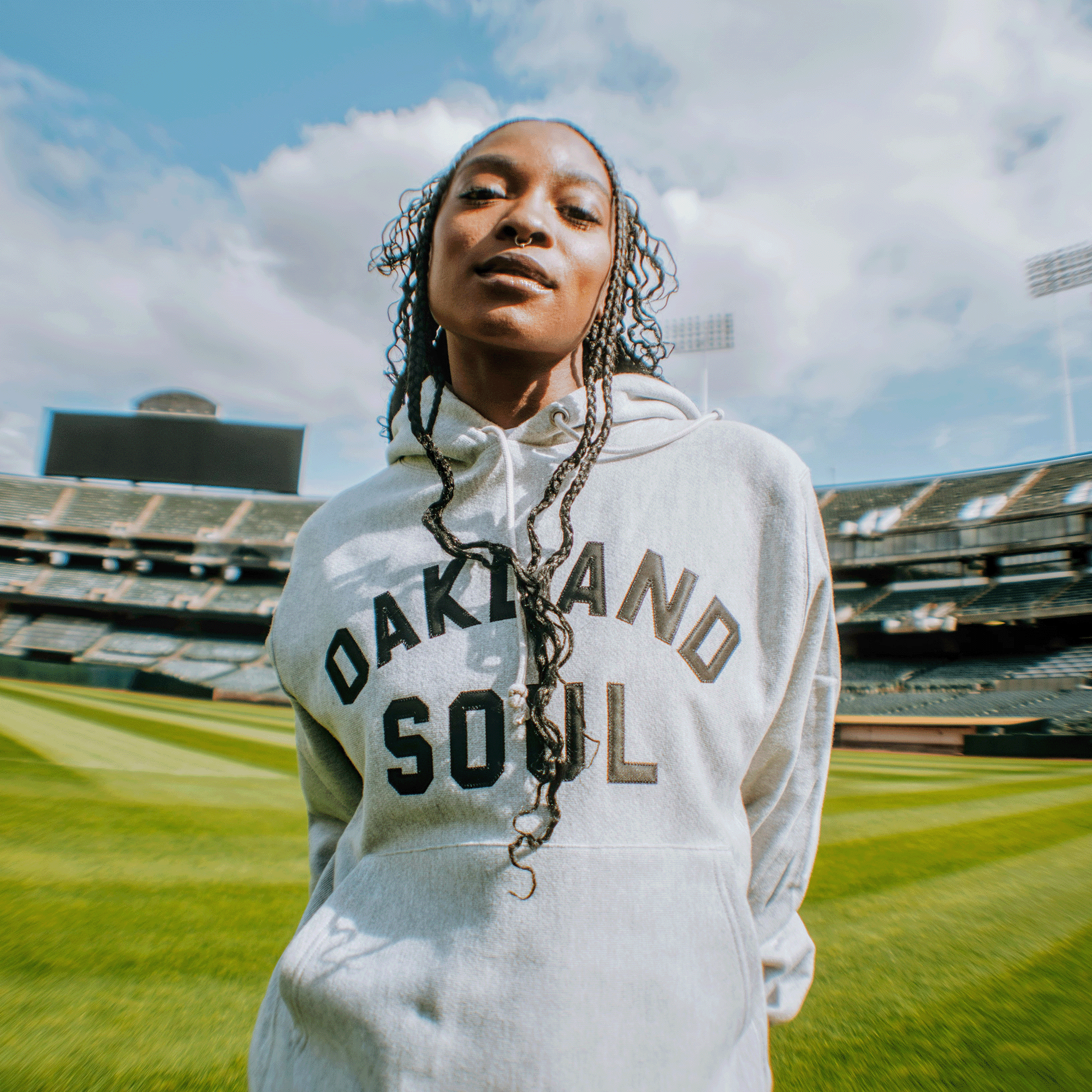 A person with long braids wears an Oakland Soul Champion X Wordmark Hoodie while standing confidently on a green sports field, with stadium seats and scoreboards in the background under a partly cloudy sky.