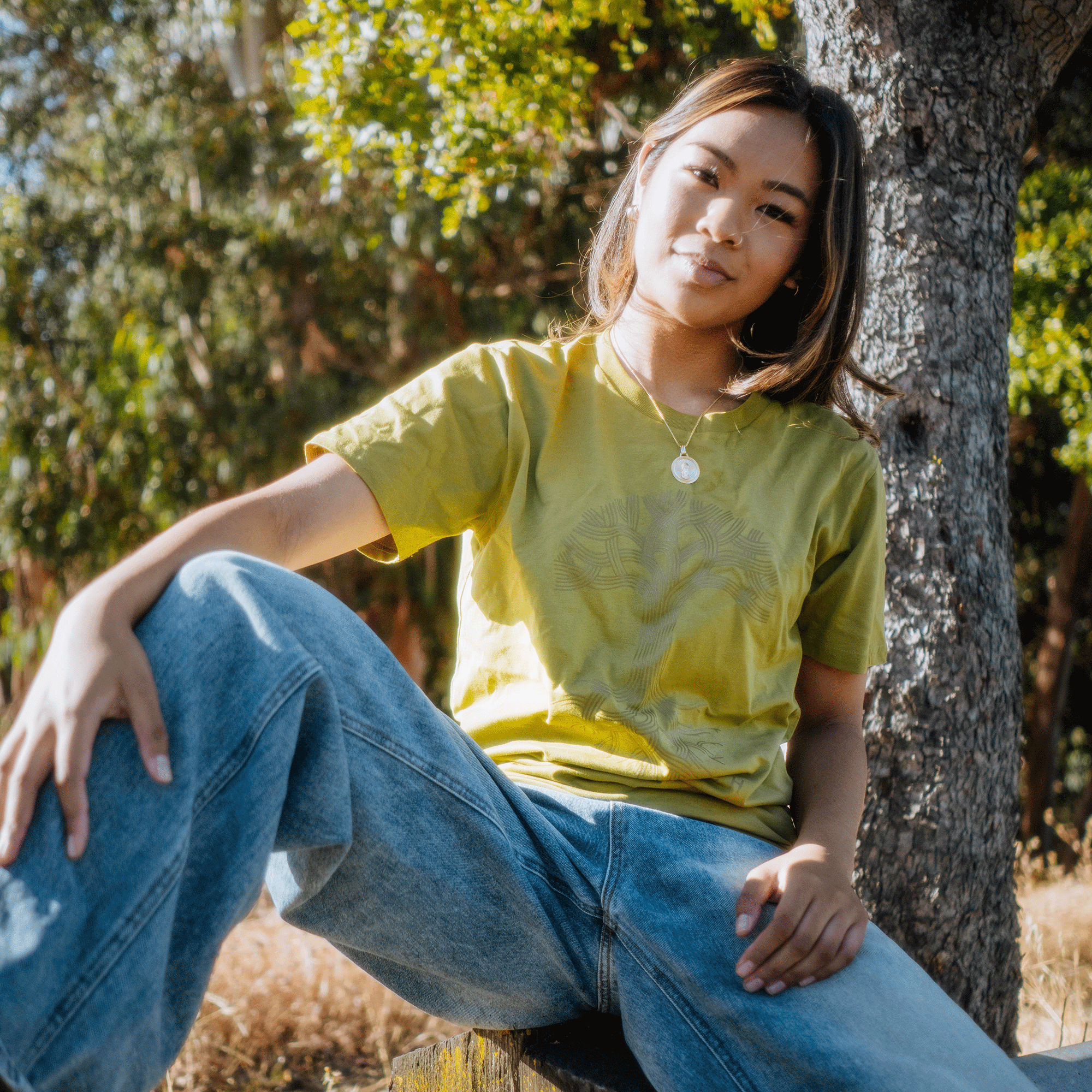 A young person sits casually outdoors on a wooden bench, wearing an Oaklandish Classic Logo Tee in yellow and blue jeans. One leg is bent and resting on the bench while the other dangles off. They are in a relaxed pose, with a classic tree and greenery in the background. Sunlight highlights their face and clothes.