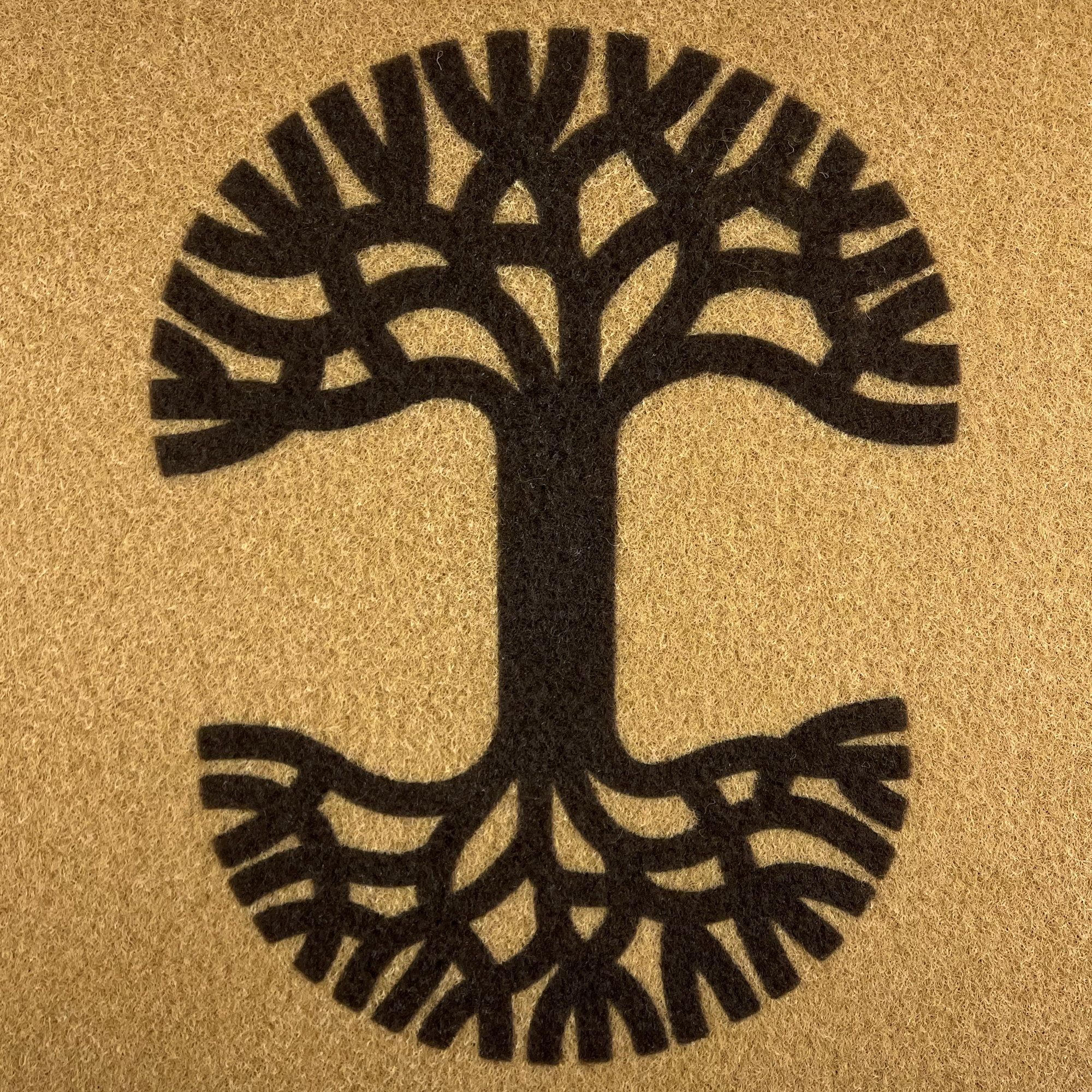 The Oaklandish Logo Door Mat by Oaklandish features a stylized tree illustration centered on a textured brown background. Perfect for both indoor and outdoor use, this door mat showcases a thick trunk with symmetrical branches and roots, forming an overall circular shape. The simple, monochromatic design in dark brown includes anti-slip backing against the lighter brown backdrop.