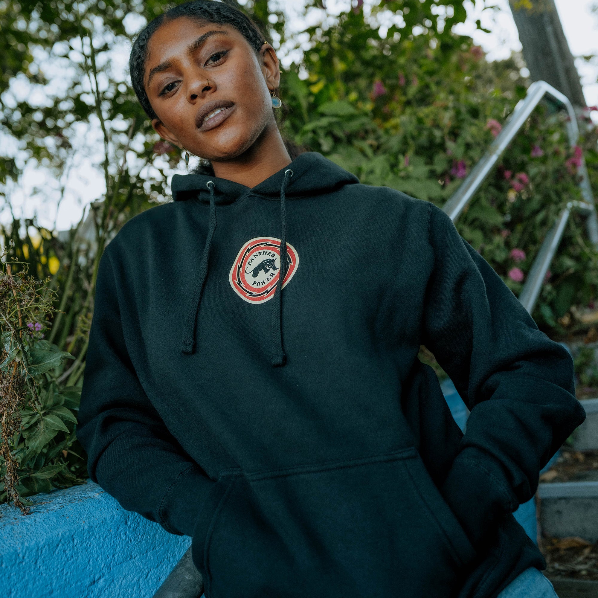 A person with dark braided hair wearing an Oaklandish Educate to Liberate Hoodie with a circular logo of Shepard Fairey's bear stands outdoors near some foliage and a blue-painted wall. They have their hands in the hoodie pocket and are looking forward with a calm expression, perfectly embodying the spirit of Educate to Liberate.