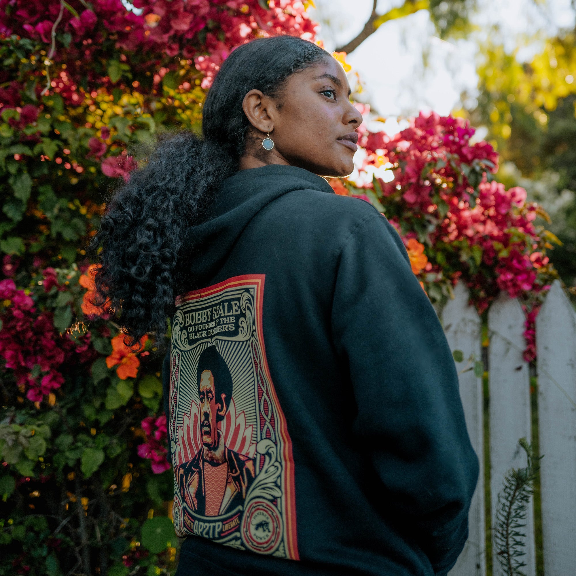 A woman with long, curly hair stands against a backdrop of vibrant pink bougainvillea. She's wearing the **Educate to Liberate Hoodie** by **Oaklandish**, featuring a colorful, artistic design by Shepard Fairey on the back. The design showcases a portrait and bold text, "Educate to Liberate." She gazes to the side, with a white picket fence and greenery in the background.