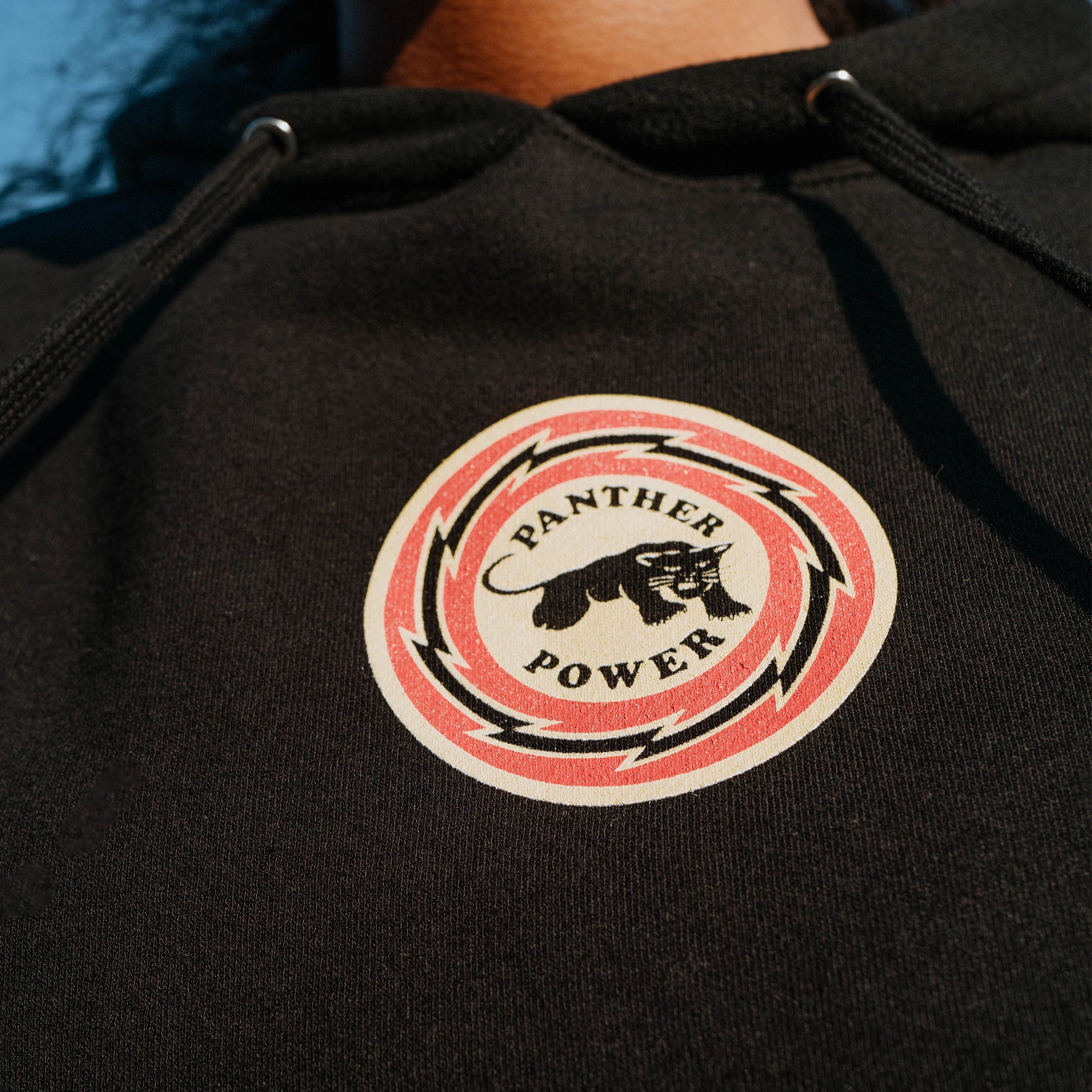 Close-up of a person wearing the Educate to Liberate Hoodie by Oaklandish. The hoodie features a round logo with a panther in the center, surrounded by a red circular design with black lightning bolts. The text "PANTHER POWER" is written around the panther in bold black font, reminiscent of Shepard Fairey's iconic style.