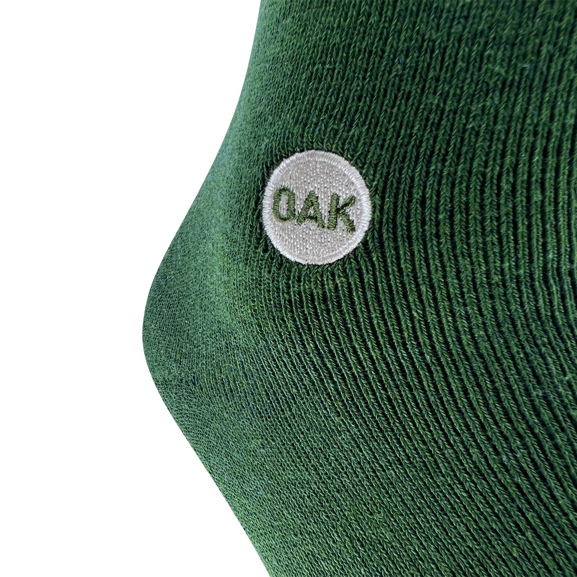 A close-up of a green fabric, likely from an Oaklandish Plush Oak Sock, with a textured, knitted pattern. There is a small white circular patch on the fabric with the uppercase letters "OAK" embroidered in green inside the patch. The background is plain white—plush socks for happy feet.
