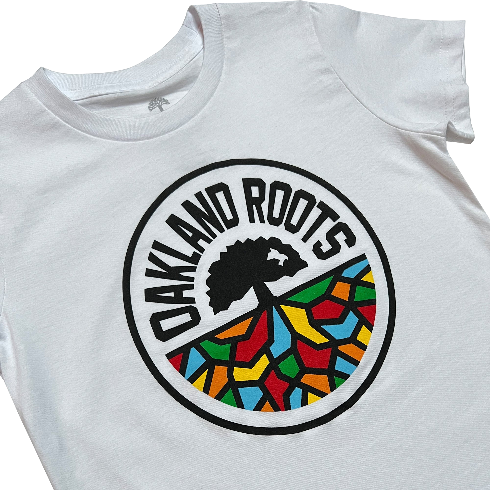 The Youth Oakland Roots SC Classic Tee by Oakland Roots SC is a white t-shirt featuring a circular emblem on the front. The emblem showcases a black tree silhouette with branching roots over a multicolored mosaic pattern, symbolizing social good. The text "OAKLAND ROOTS," representing the Oakland sports club, is printed in all caps around the top of the circle.