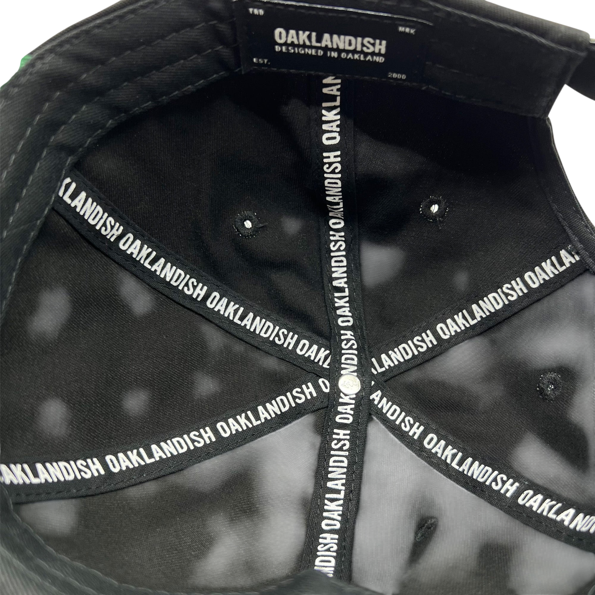 The interior view of the Official Micro Logo Dad Hat in black showcases the brand "Oaklandish" printed repeatedly along the inner seams. A label on the inside reads "Oaklandish Designed in Oakland Est. 2000." The cap features an embroidered "O" and multiple triangular sections converging at the top button.