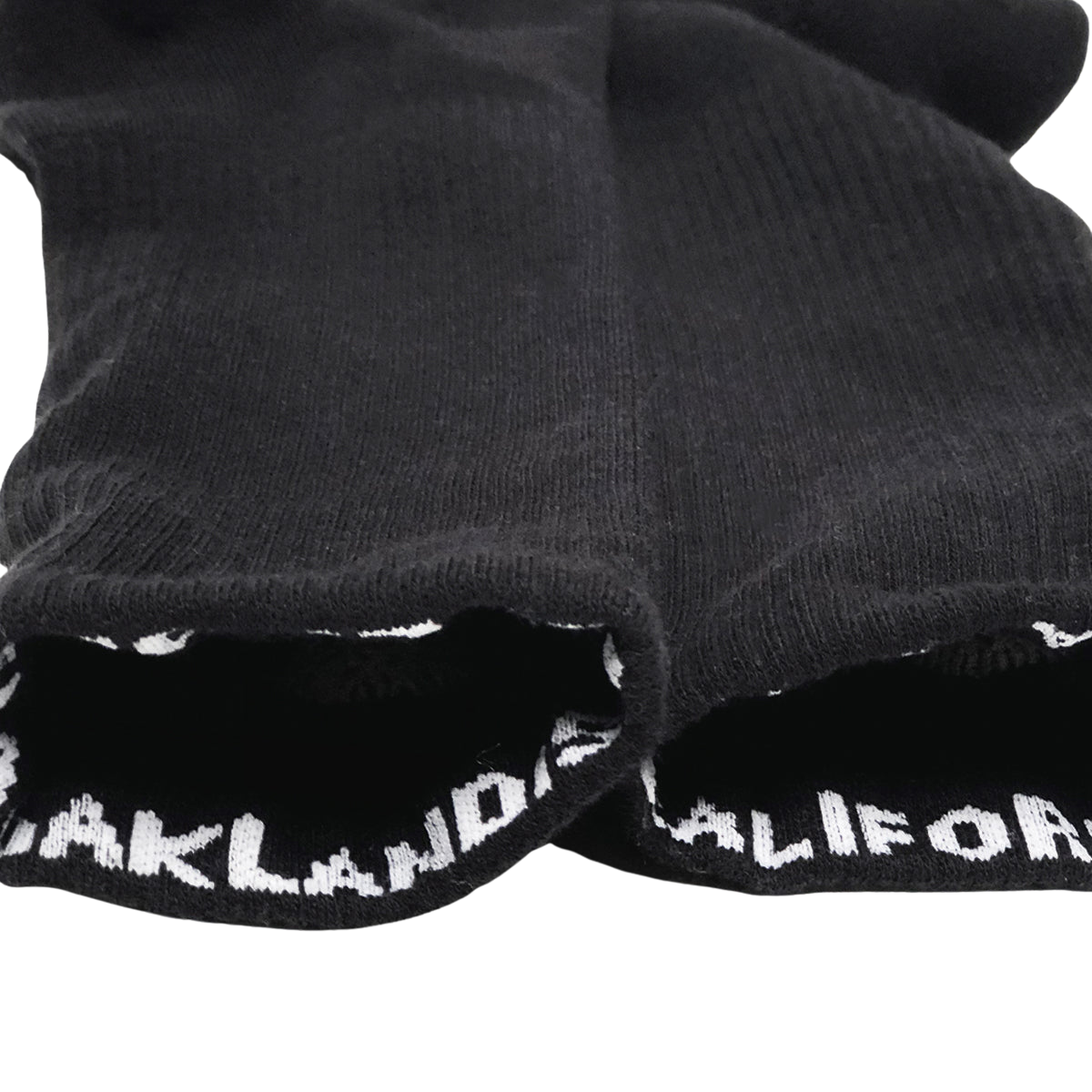 A pair of black Embroidered Logo Socks from Oaklandish is laid flat on a white surface. The cuff of each sock features the text "OAKLAND CALIFORNIA" in white, capital letters. The socks' fabric texture and knitted structure are visible, designed for adult sizes with a stylish nod to the brand's signature aesthetic.