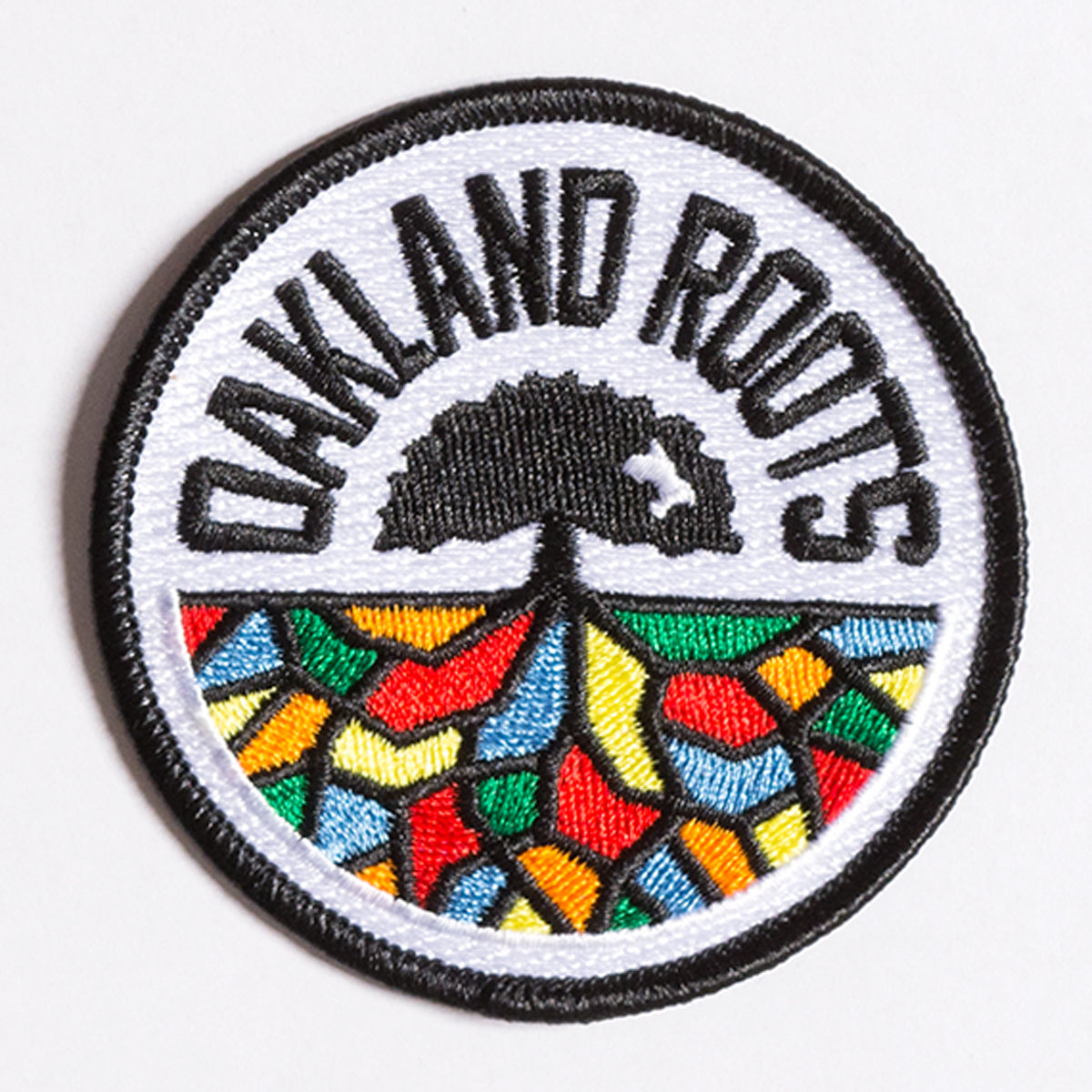 The Oakland Roots SC Patch, crafted by Oakland Roots SC, is a circular embroidered iron-on accessory featuring a black-bordered white background. "OAKLAND ROOTS" is inscribed in black at the top, framing an image of a tree. Beneath the tree, the roots are depicted as a vibrant mosaic with segments in red, orange, yellow, green, and blue—ideal for fans of the Oakland Roots Sports Club.