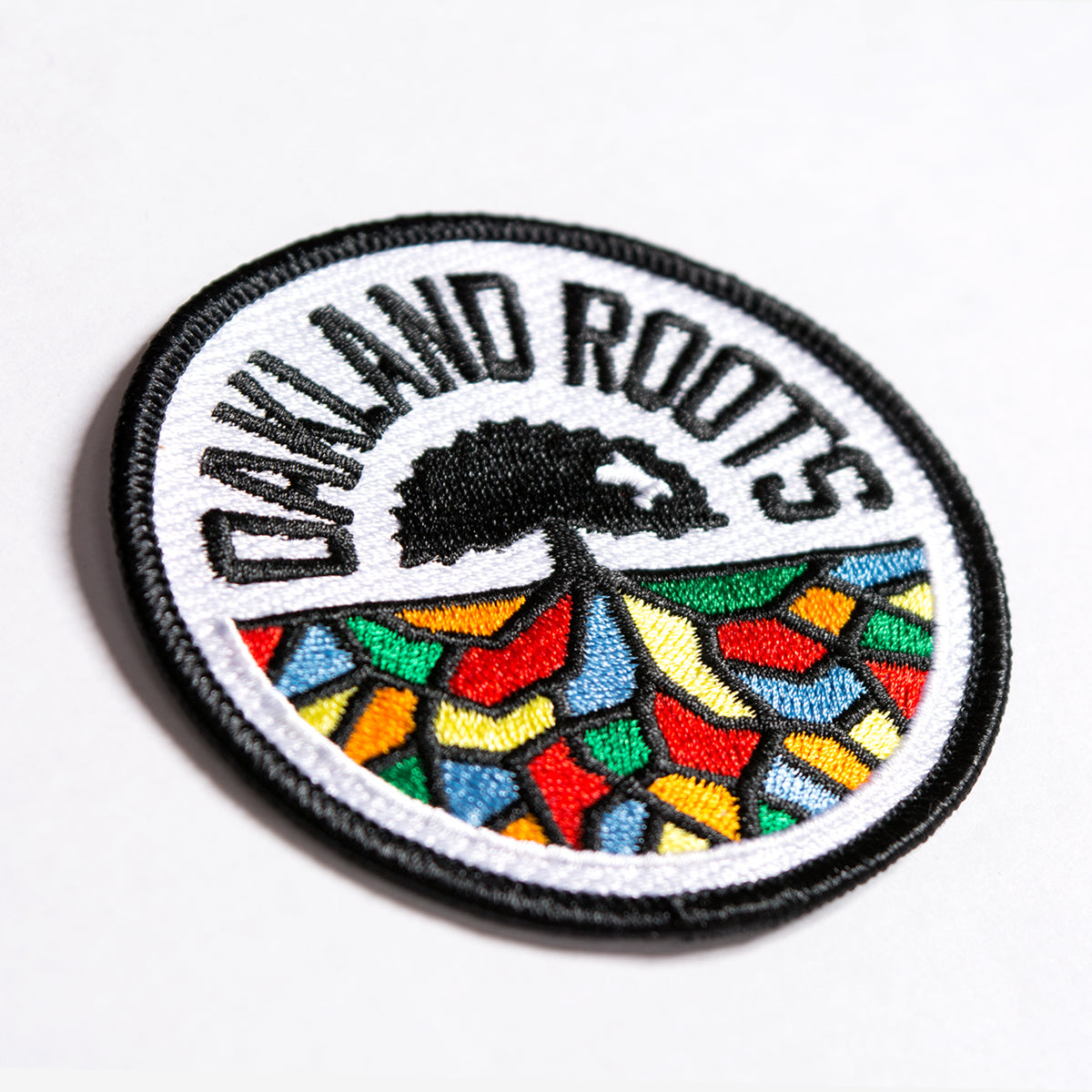 Get your hands on the Oakland Roots SC Patch by Oakland Roots SC, a circular embroidered iron-on patch featuring a black border and "OAKLAND ROOTS" text in black at the top. The center showcases a black tree with roots extending over a vibrant mosaic of red, blue, yellow, and green geometric shapes on a white background—ideal for any Oakland Roots Sports Club fan.