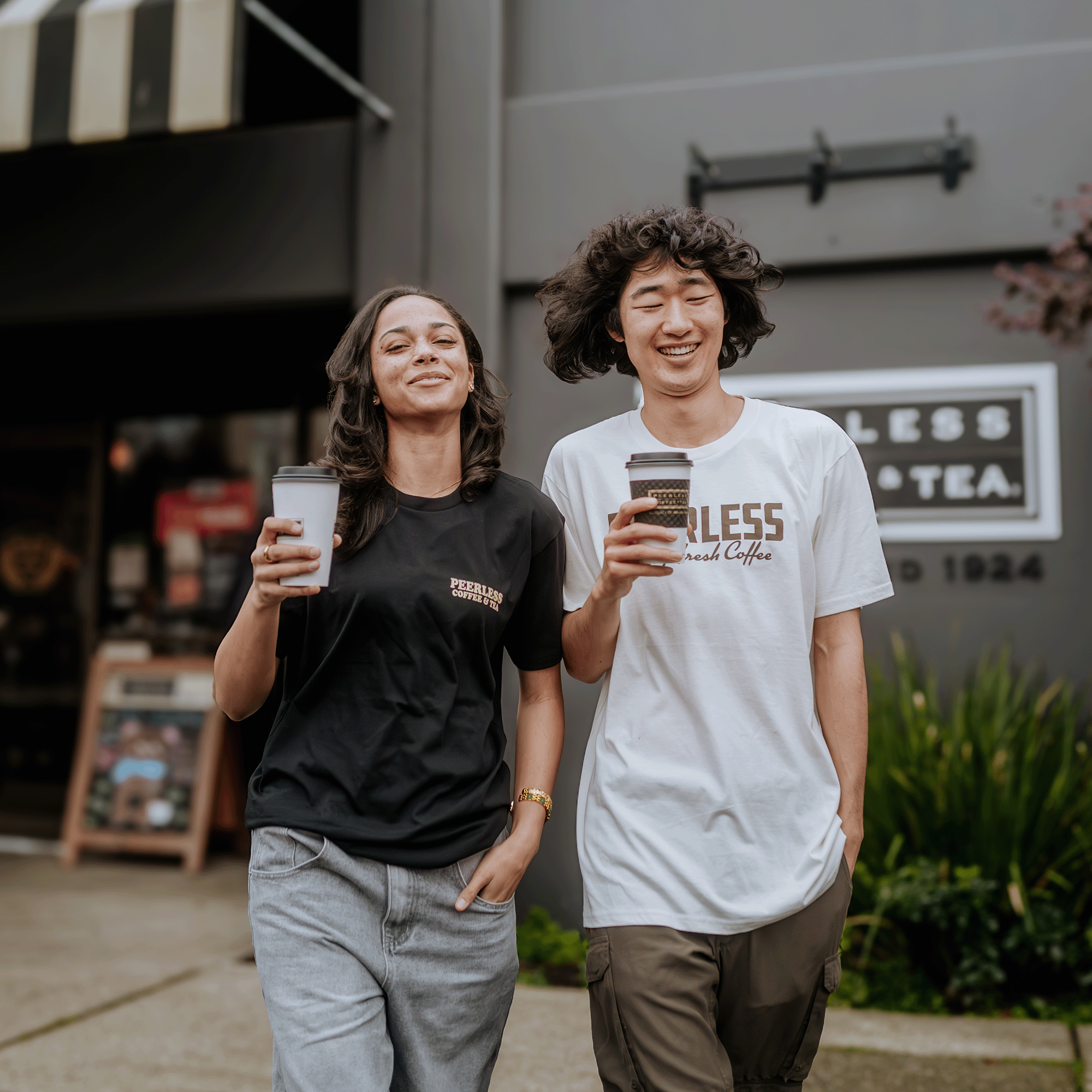 A man and a woman enjoy takeaway coffee as they walk. She wears a black t-shirt and jeans, while he sports an Oaklandish Peerless Skyline Tee and olive pants. They stroll outside a cafe with blurred signage, exuding relaxation amidst the vibrant greenery of the Oakland cityscape.