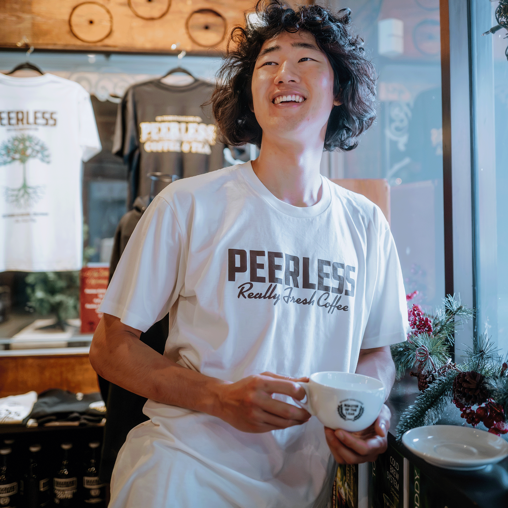 A curly-haired person smiles while holding a white "Peerless Really Fresh Coffee" cup, wearing an Oaklandish Peerless Arabica Tee. Matching tees and bottles are behind them, with a small holiday decoration adding festivity on the right.