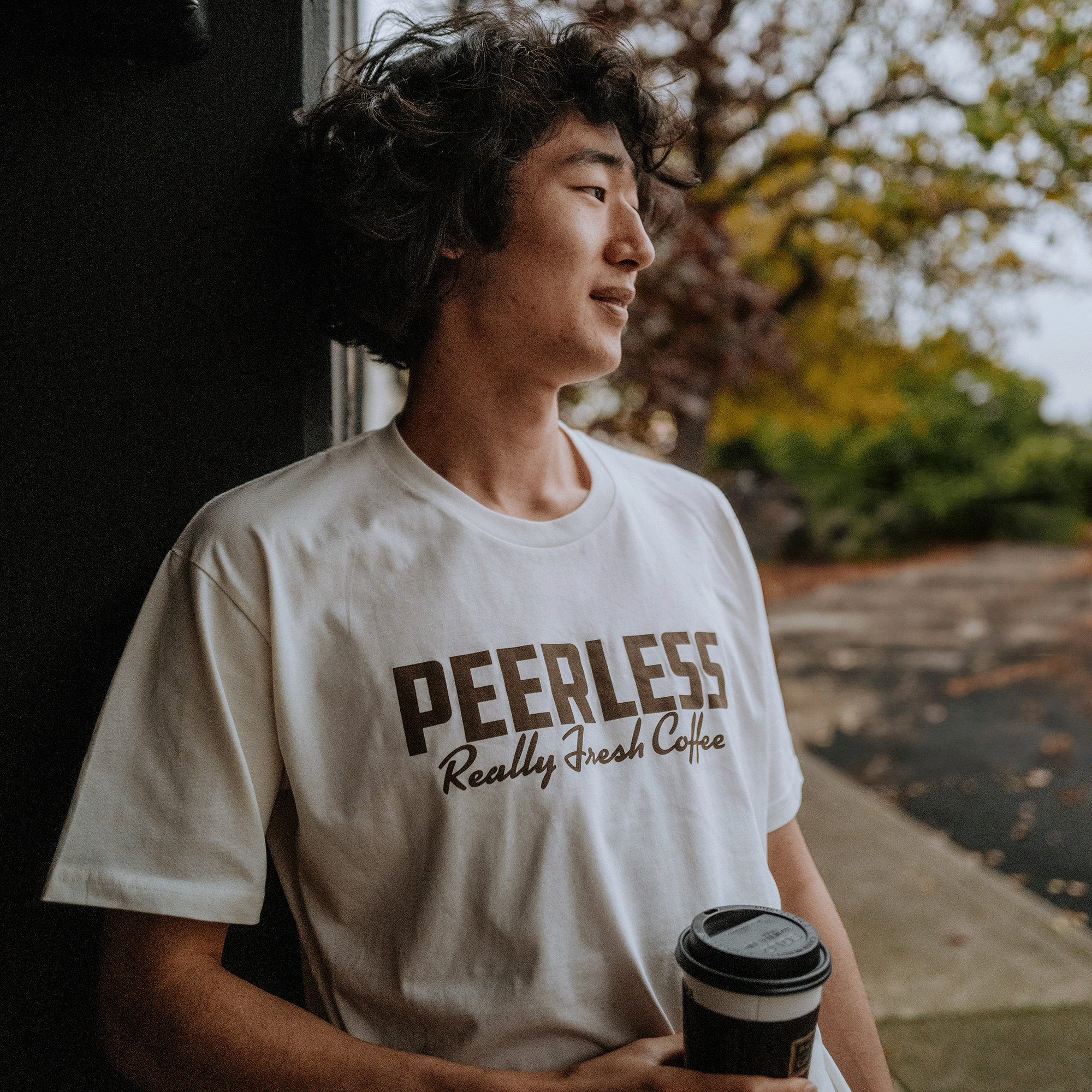 A young man with wavy hair leans against a wall, holding a paper cup. His white T-shirt reads "Peerless Arabica Tee" by Oaklandish, suggesting its link to Arabica coffee trees. He gazes into the distance, framed by trees and a path on this cloudy day.