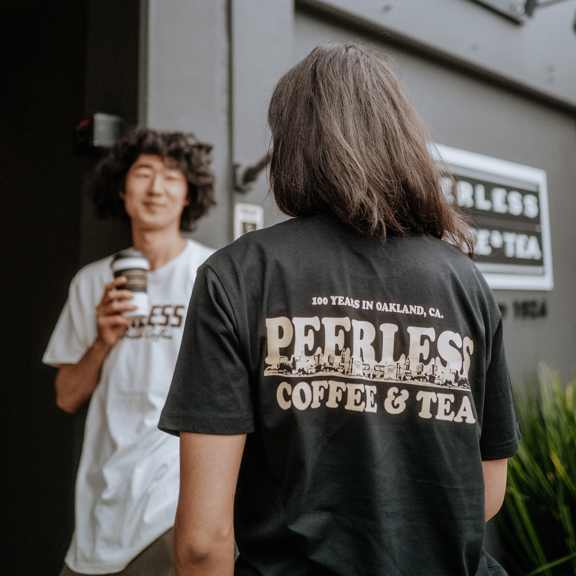 A person with curly hair holds a coffee cup, wearing the white "Peerless Coffee & Tea" shirt. Next to them, someone in black wears an "Oaklandish Peerless Skyline Tee," featuring the text "100 Years in Oakland, CA." They stand outside under the iconic sign against vibrant Oakland cityscape.