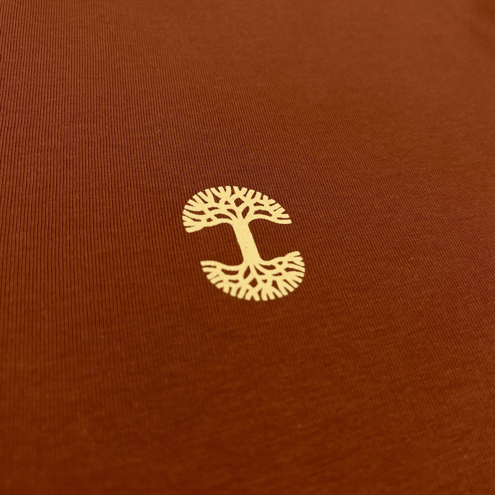 The Peyo Tee, a product by Oaklandish, features a minimalist logo of a white tree with symmetrical branches and roots in the shape of an "S," centered on a rust-colored fabric background. This intricate design is showcased on a classic fit t-shirt worn by a female model, highlighting clear lines representing the tree's structure.