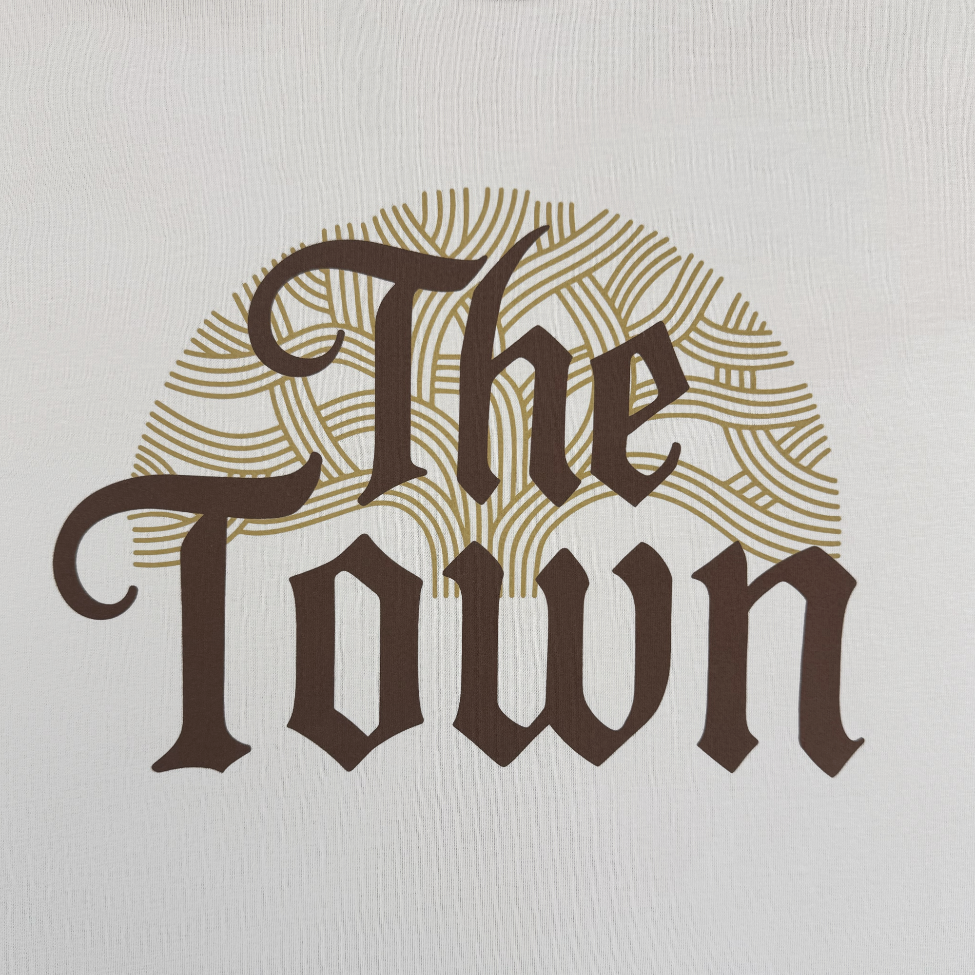The image displays "The Town" in a bold, classic serif font over a gold, wavy semi-circle—reflecting Oaklandish style—on a white background. This design is featured on the GSW Pirate Black-Letter LS Tee from the Town Collection.