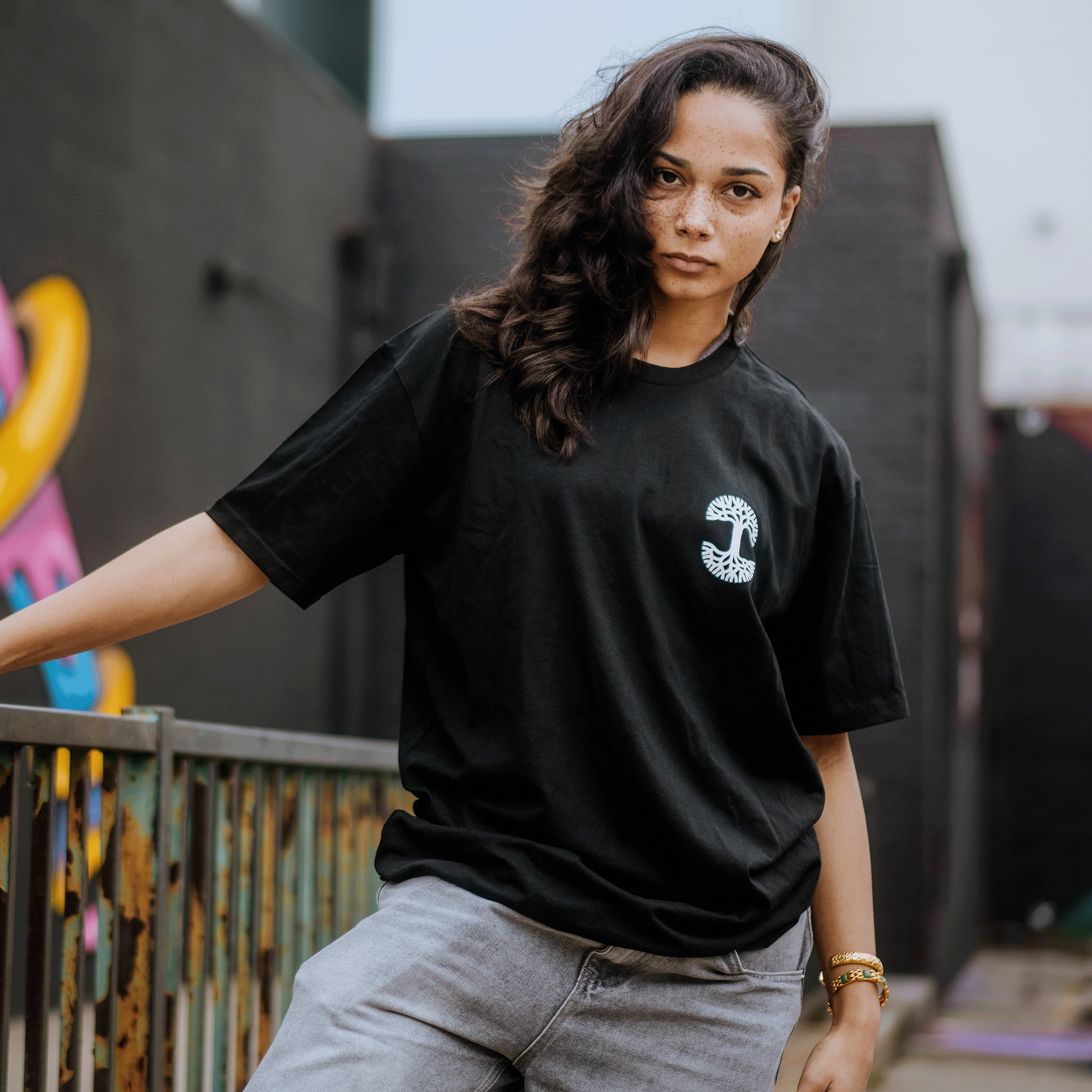 A person with long, dark hair leans against a railing outdoors wearing the Posy Tee from Oaklandish. The black cotton shirt features a subtle design and is paired with gray pants. They stand against a graffiti-adorned black wall, looking at the camera calmly.