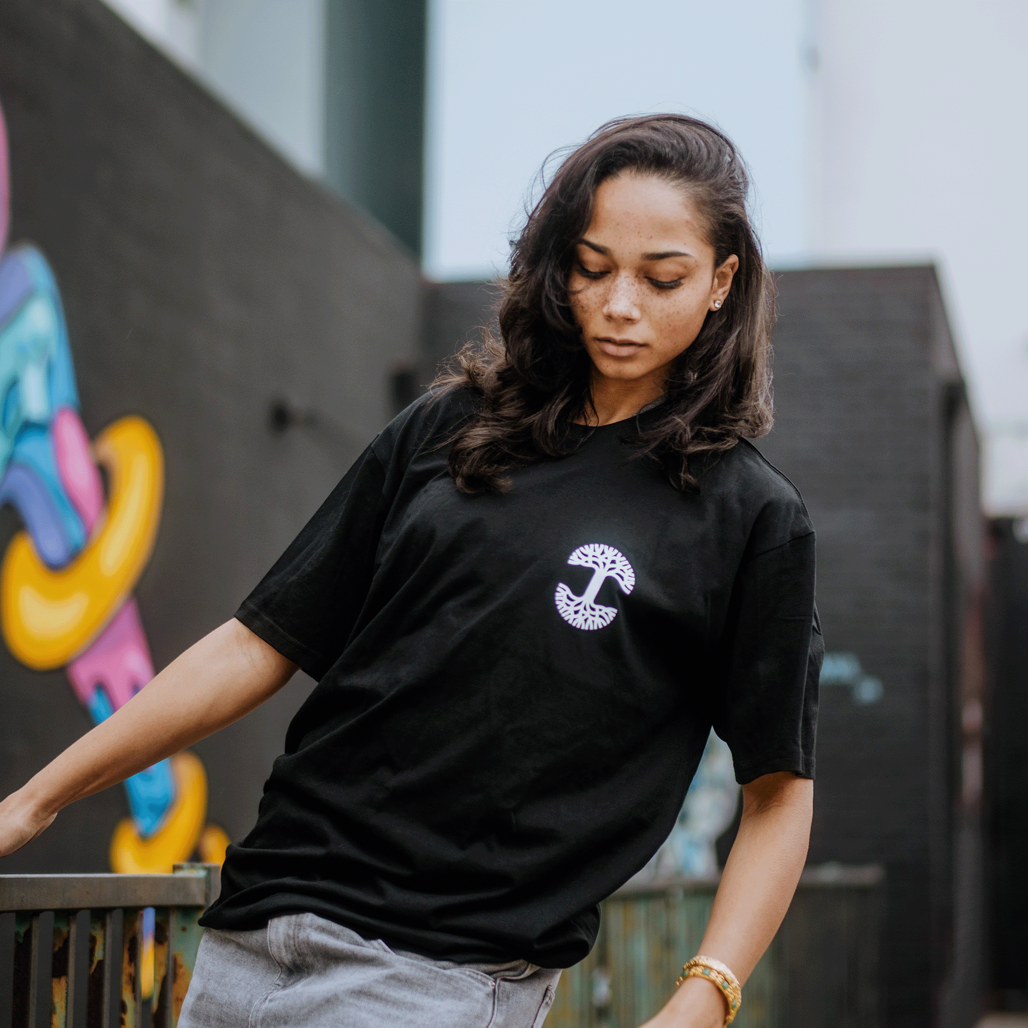 A long-haired woman in a classic fit, black "Posy Tee" by Oaklandish displays a chest graphic as she stands by an outdoor railing. With a calm gaze directed downward, a vibrant graffiti-covered wall serves as her backdrop.