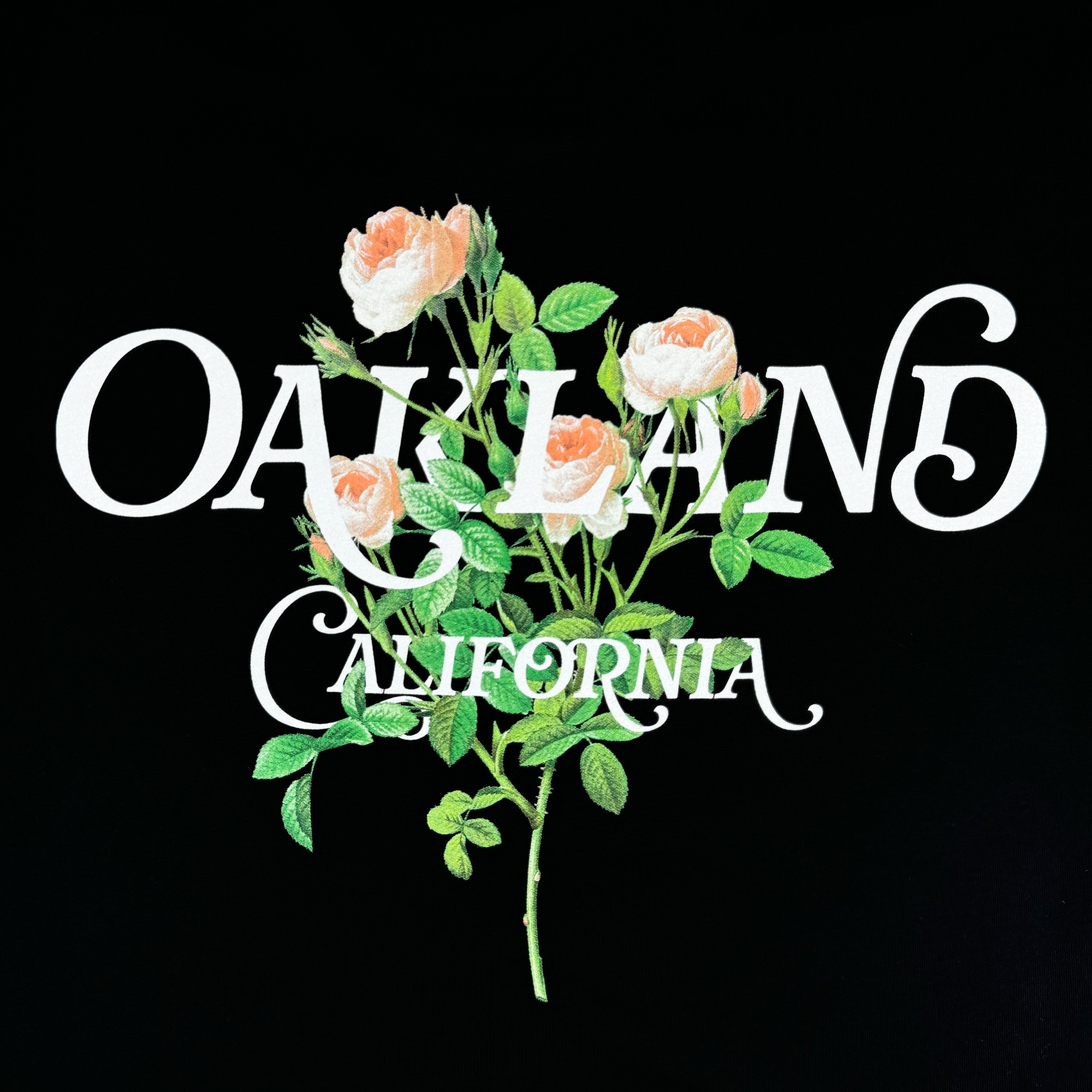 The Posy Tee by Oaklandish features the words "OAKLAND CALIFORNIA" in white script against a black backdrop, ideal for a stylish men's cotton t-shirt. A branch adorned with delicate white and pale pink roses and green leaves intersects the text, adding a subtle floral accent.