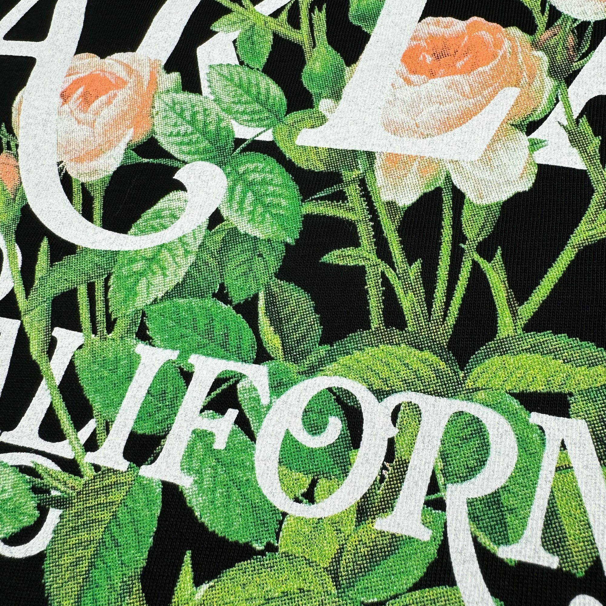 A detailed view of the cotton fabric on the Posy Tee by Oaklandish highlights a floral pattern featuring pink roses and green leaves, interwoven with bold white "California" lettering against a black background. Ideal for a men's collection t-shirt, its texture and vibrant colors are strikingly prominent.