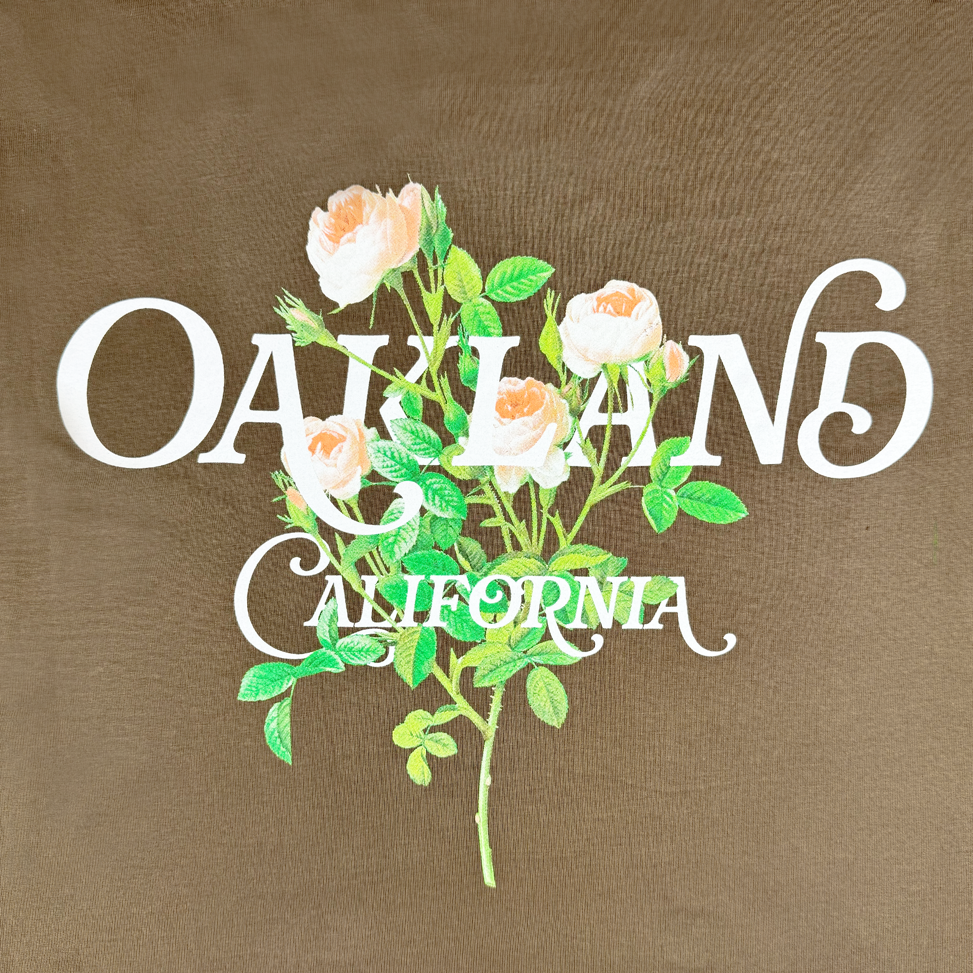 Introducing the Posy Tee by Oaklandish, a cotton t-shirt featuring bold white "OAKLAND" lettering with "California" elegantly scripted below. An eye-catching backdrop of white-petaled roses with green leaves on a brown background makes this a perfect addition to your wardrobe collection.