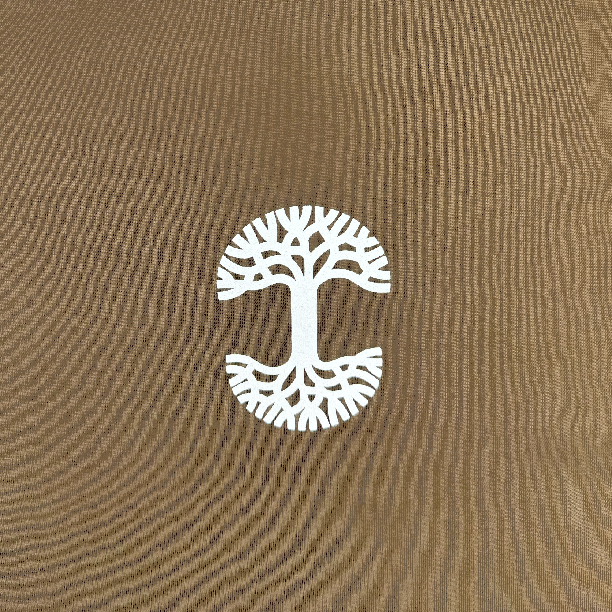 The Posy Tee from Oaklandish features a white, stylized tree with outstretched branches and roots on the front of a brown cotton t-shirt. The circular design, where the branches mirror the roots in symmetry, creates an eye-catching pattern that's part of our latest collection, complemented by a textured background that adds subtle contrast.