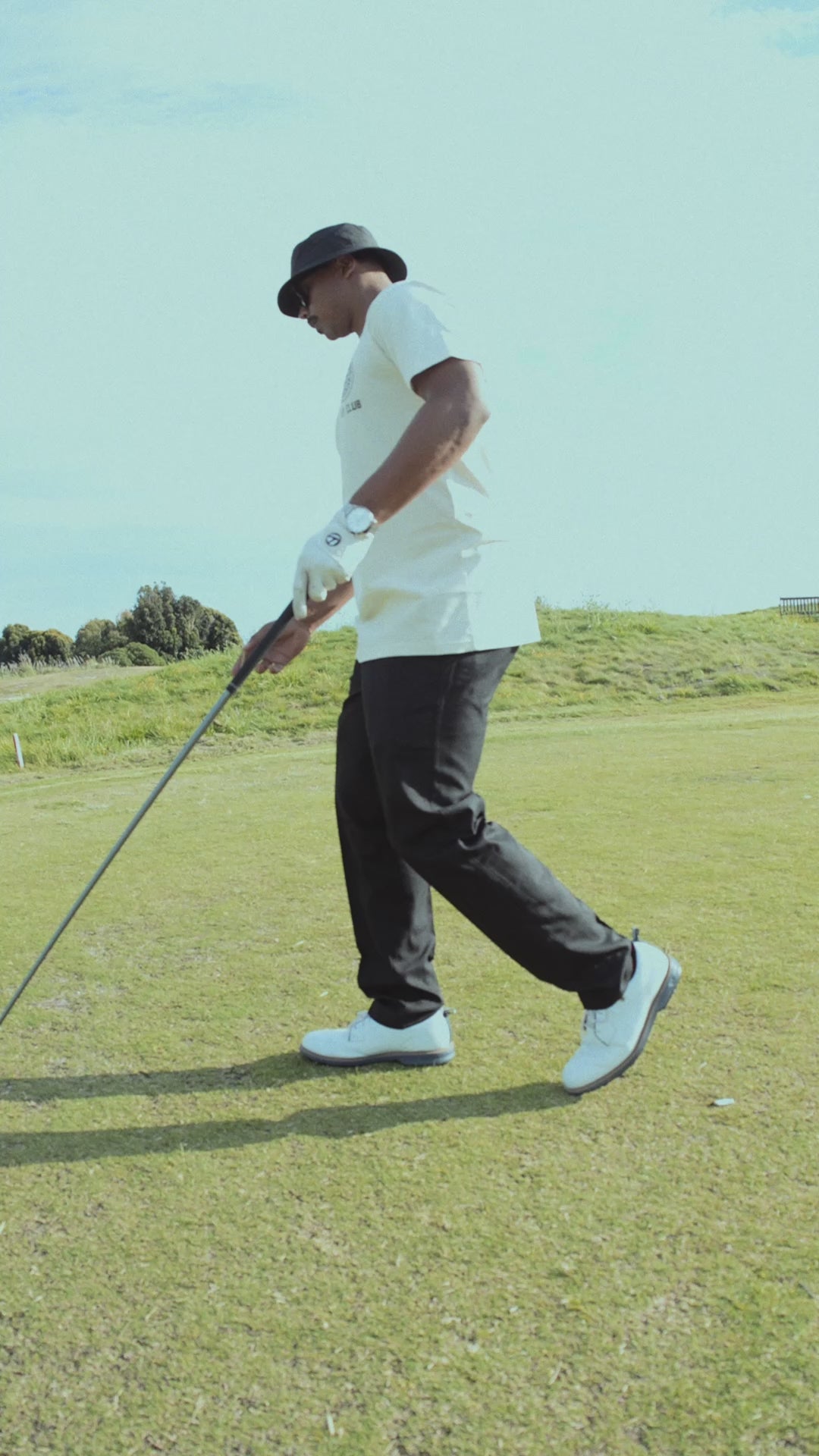 Guy in bucket hat on golf course swings club.