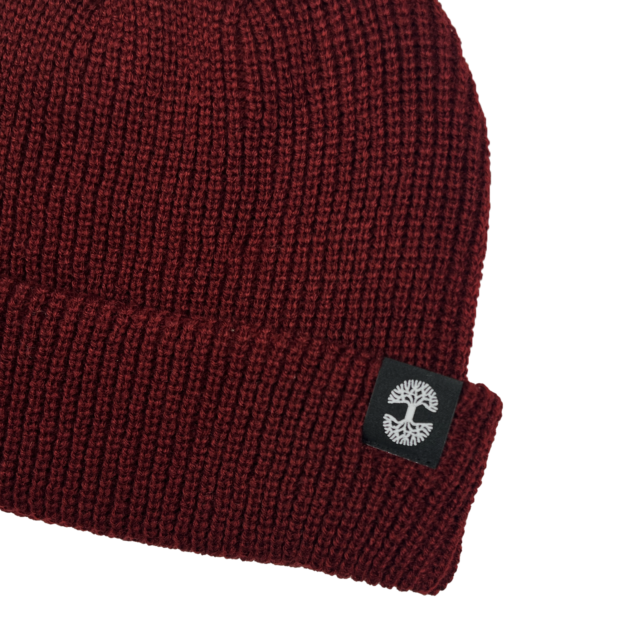 A close-up of the Ramos Beanie by Oaklandish, a dark red knit crafted from woven acrylic with a folded brim. The brim showcases a black patch featuring a white abstract tree logo on the side. This stylish piece of headwear has a textured, ribbed pattern and is set against a plain, light gray background.