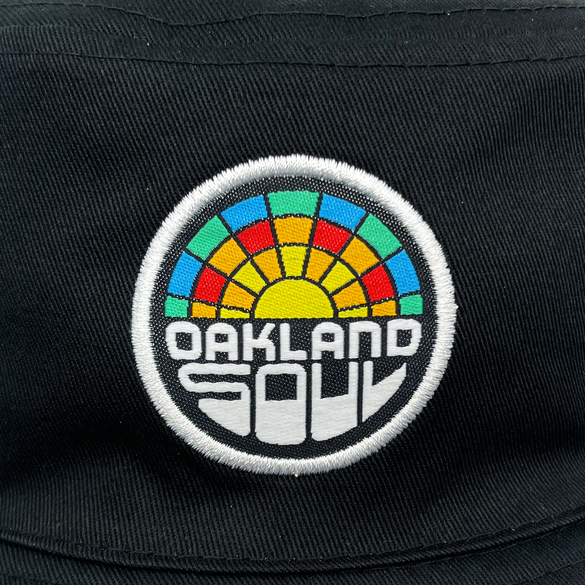 A circular patch is embroidered on a black fabric background featuring a vibrant sunburst design with segments in red, yellow, green, and blue. Below the sunburst, "OAKLAND SOUL" is stitched in bold, white geometric letters. This striking patch will look perfect on the Oakland Roots SC Reversible Bucket hat from the Oakland Roots SC brand.