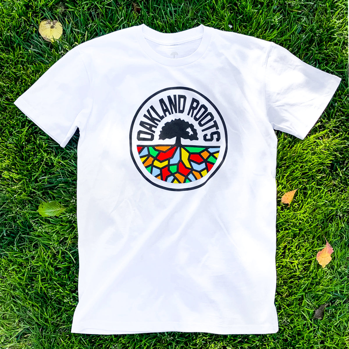 A white Oakland Roots SC Classic Tee lies on grass, showcasing the distinct "OAKLAND ROOTS" circular logo in its center. The logo features a black tree with roots extending into a vibrant mosaic of red, yellow, green, and blue shapes beneath it. Brown and yellow leaves are scattered on the grass around the tee, representing the blend of sports and social good that the Oakland Roots SC embodies.