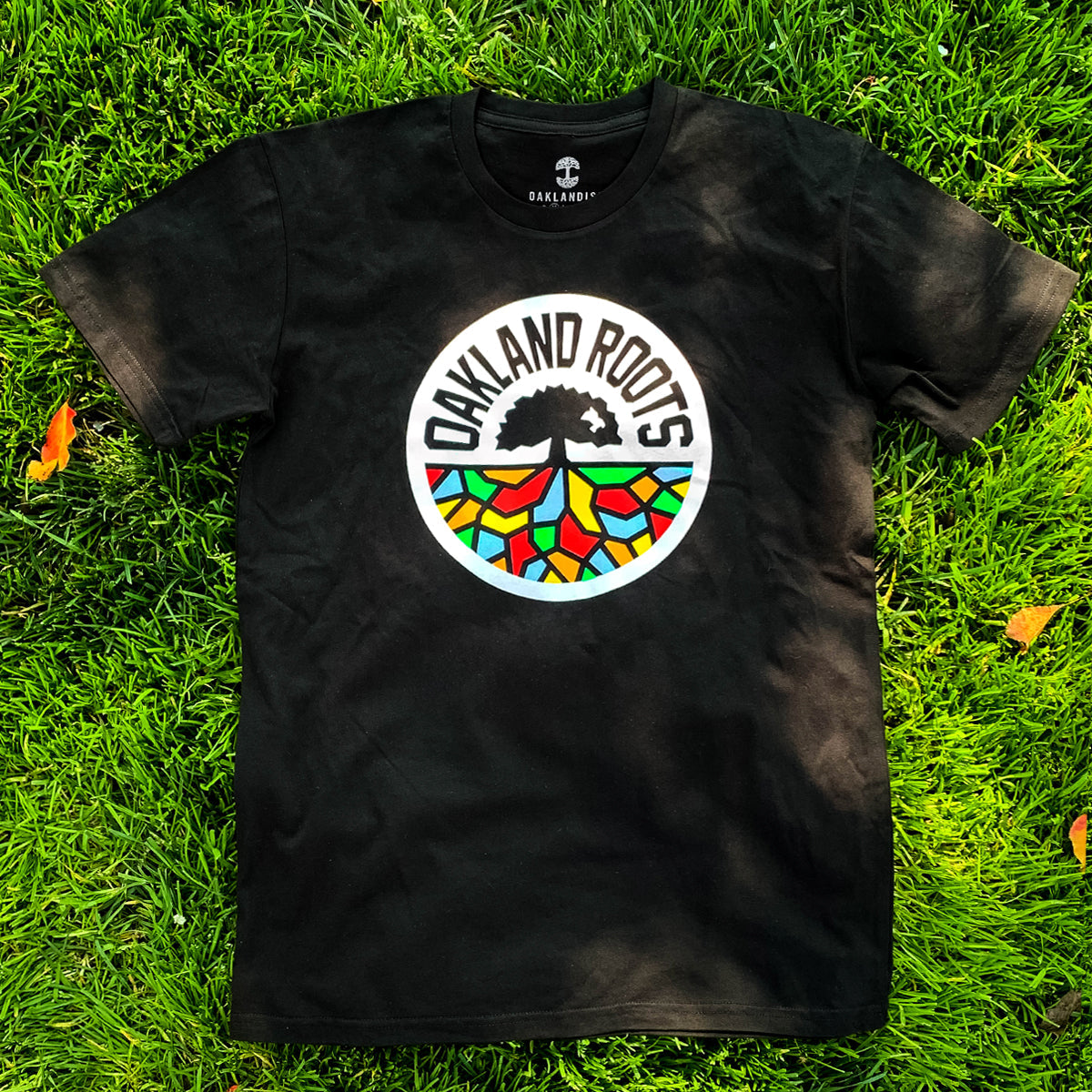 An Oakland Roots SC Classic Tee lies on vibrant green grass. The black T-shirt features a colorful circular logo in the center that reads "Oakland Roots Sports Club" around the top, displaying a tree with multicolored roots extending downward, symbolizing social good and community connection.