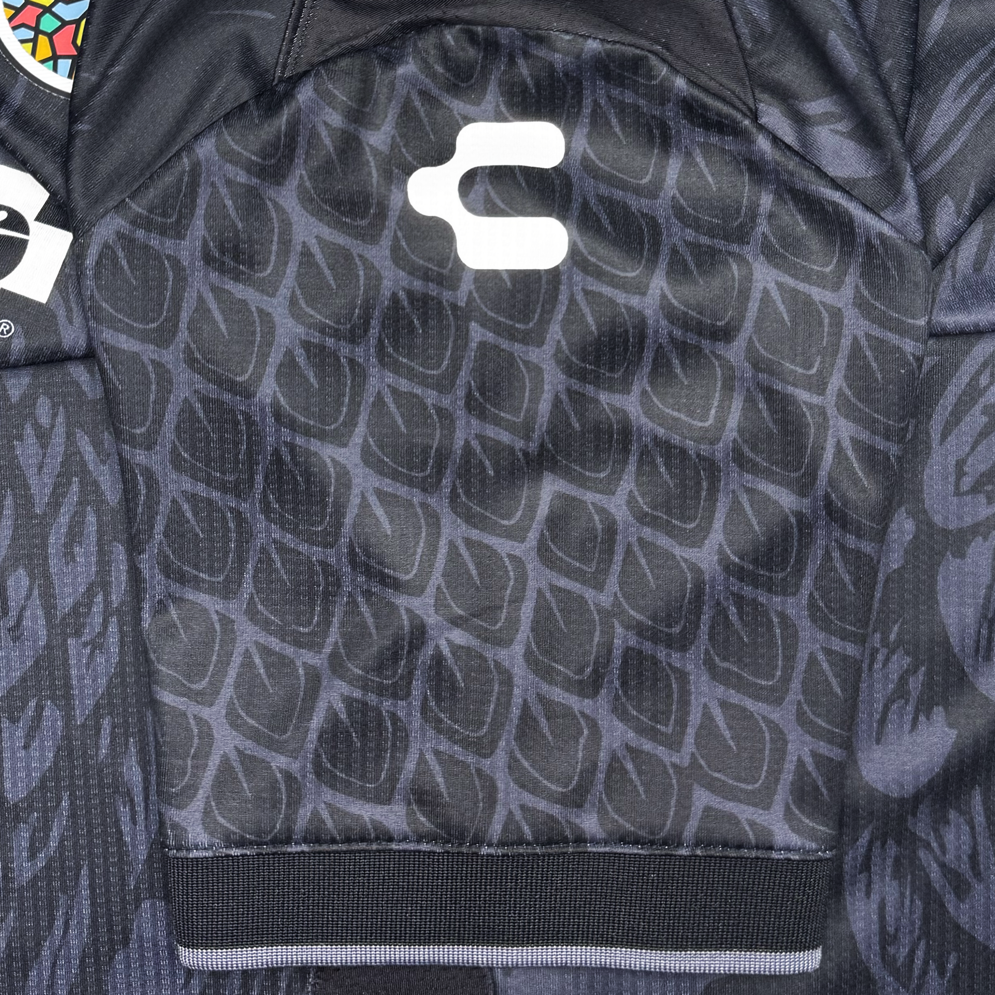 Close-up of the Oakland Roots SC Custom Unisex Oak-ness Awakens Kit: a black jersey featuring a subtle geometric scale-like pattern inspired by Oak-ness Monster folklore, a white logo within a colorful circular badge, and a black ribbed bottom cuff.
