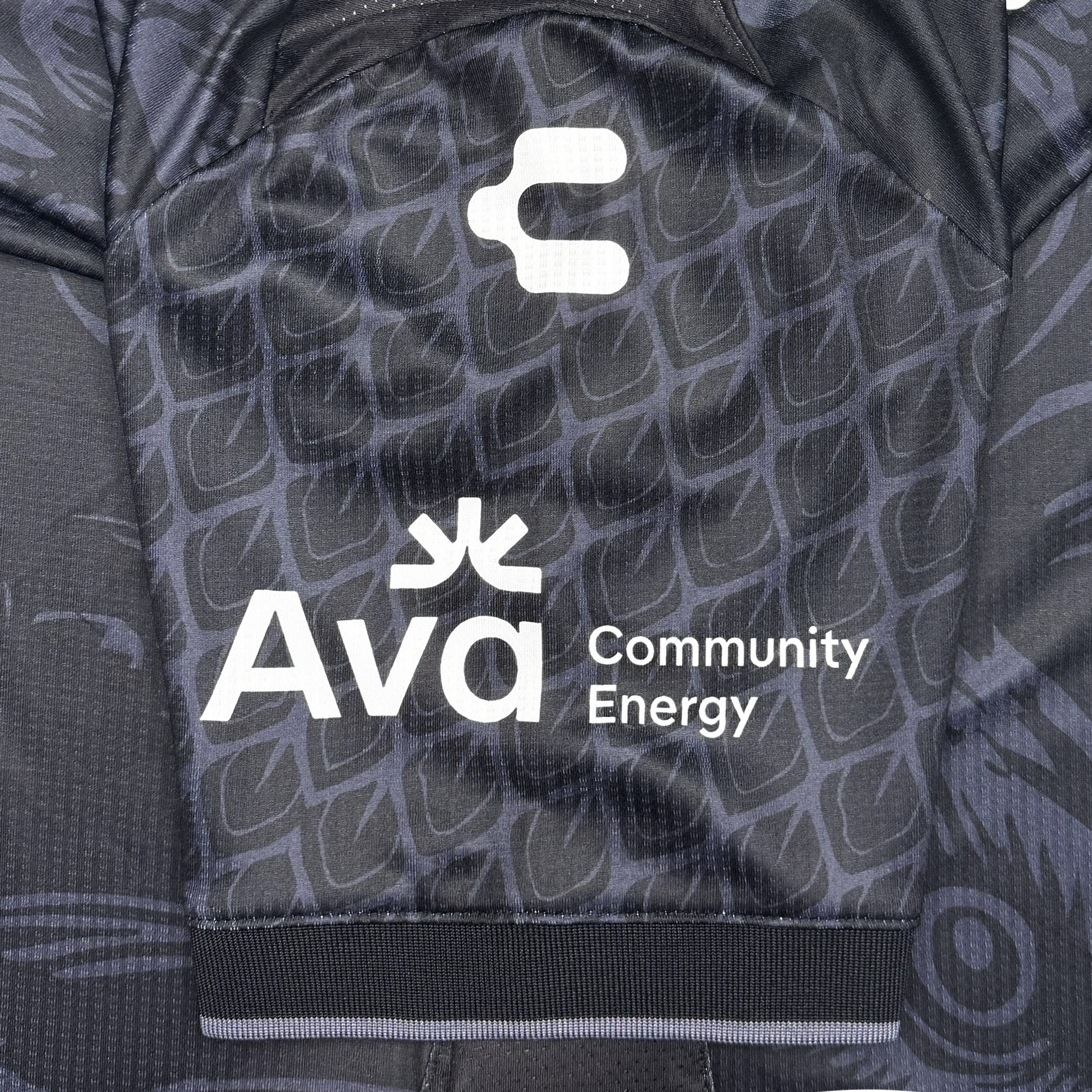Close-up of the Unisex Oak-ness Awakens Kit by Oakland Roots SC: a black sports jersey with subtle geometric patterns. The sleeve displays white logos, including “Ava Community Energy” and an abstract icon, popping against the dark background.