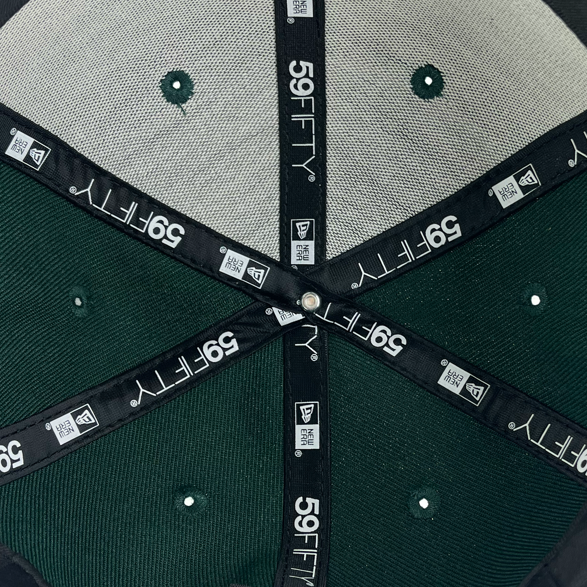 A close-up view of the inside of a New Era Oakland Roots SC 59FIFTY cap reveals interior seams reinforced with black fabric tape featuring the 59FIFTY logo, New Era logo, and additional branding text. The green and gray panels with ventilation holes make this Oakland Roots SC-inspired cap a standout piece.