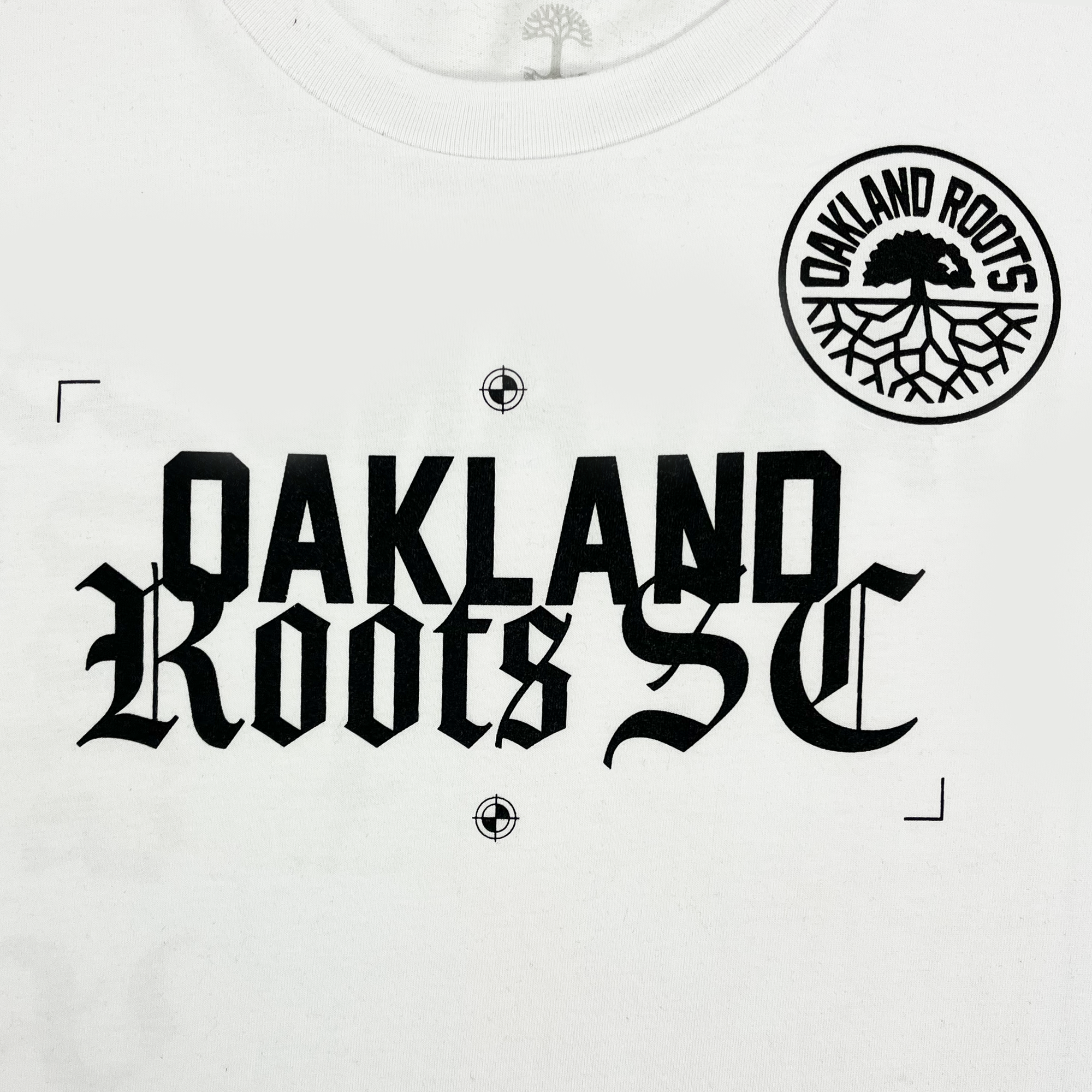 The Oakland Roots SC Futbol Town Tee is a white T-shirt that prominently displays the text "OAKLAND Roots SC" in bold, black gothic font across the chest. Additionally, there is a circular logo above the text on the left side, featuring the words "Oakland Roots Sports Club" encircling a tree with roots.