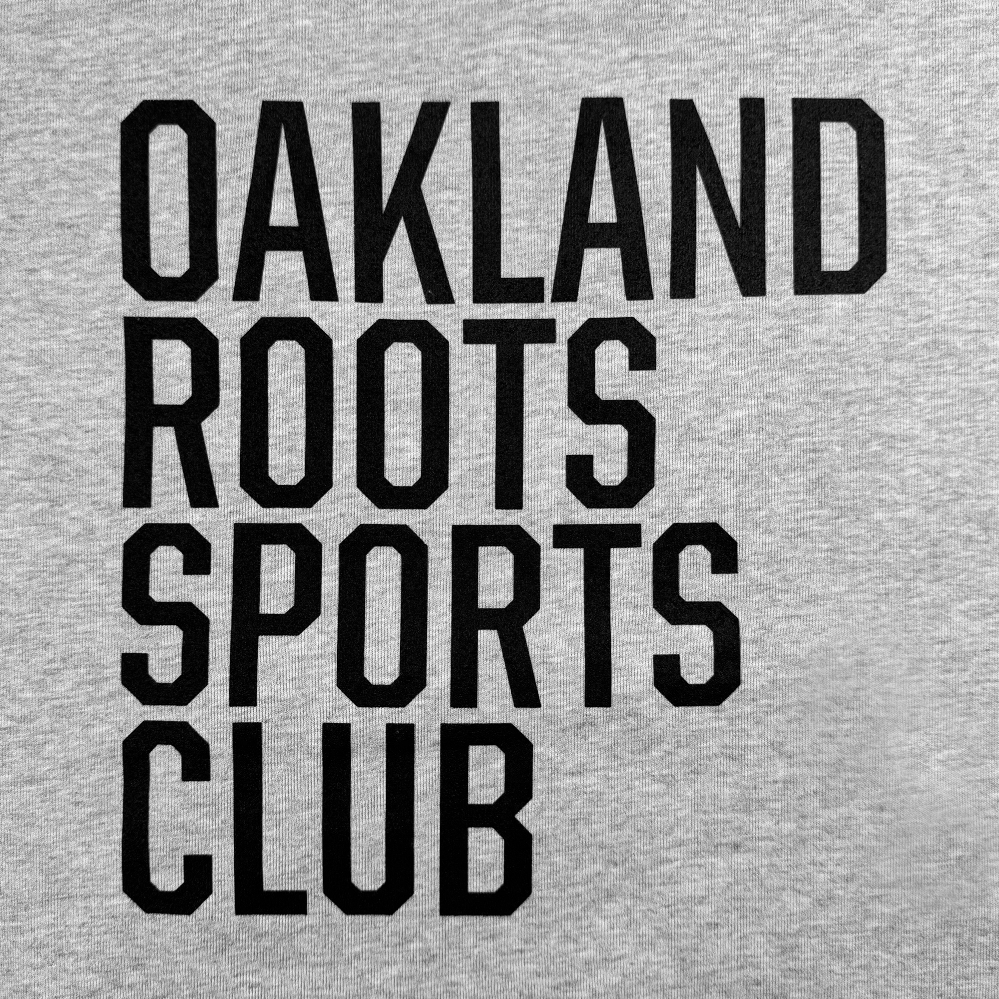 The image displays the text "OAKLAND ROOTS SPORTS CLUB" in bold, black, uppercase letters across three lines on a light gray background. This design on the Oakland Roots SC Heavy Hoodie by Oakland Roots SC highlights the club's dedication to social good and community impact with a compelling visual contrast.