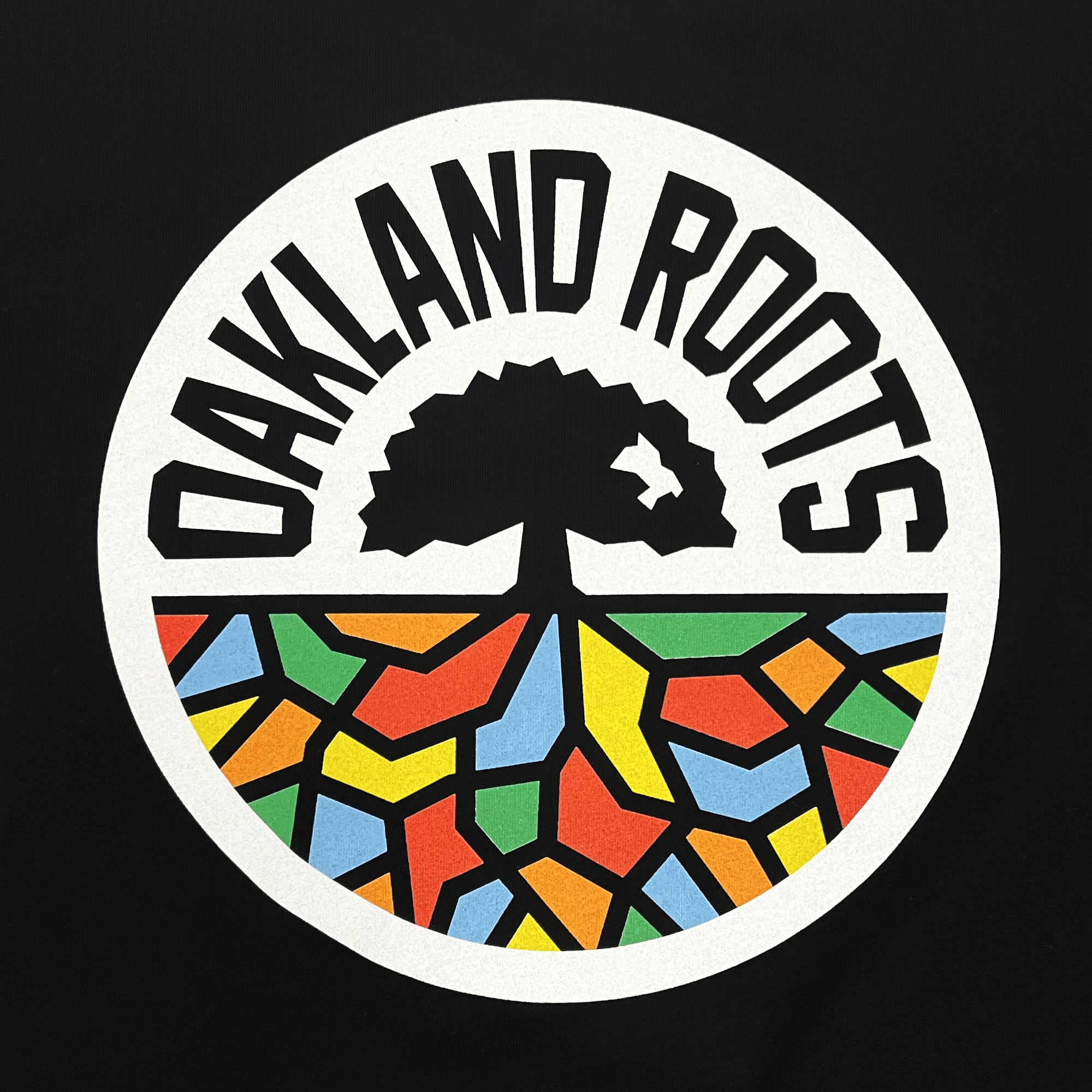 The circular logo on the Women's Oakland Roots SC Classic Relaxed Tee from Oakland Roots SC, perfect for a t-shirt, is set against a black background. The top half showcases bold, black capital letters spelling "OAKLAND ROOTS," curving around an intricately branched tree. Below, a vibrant mosaic of geometric shapes in red, yellow, blue, and green encapsulates the dynamic essence of this sports club.