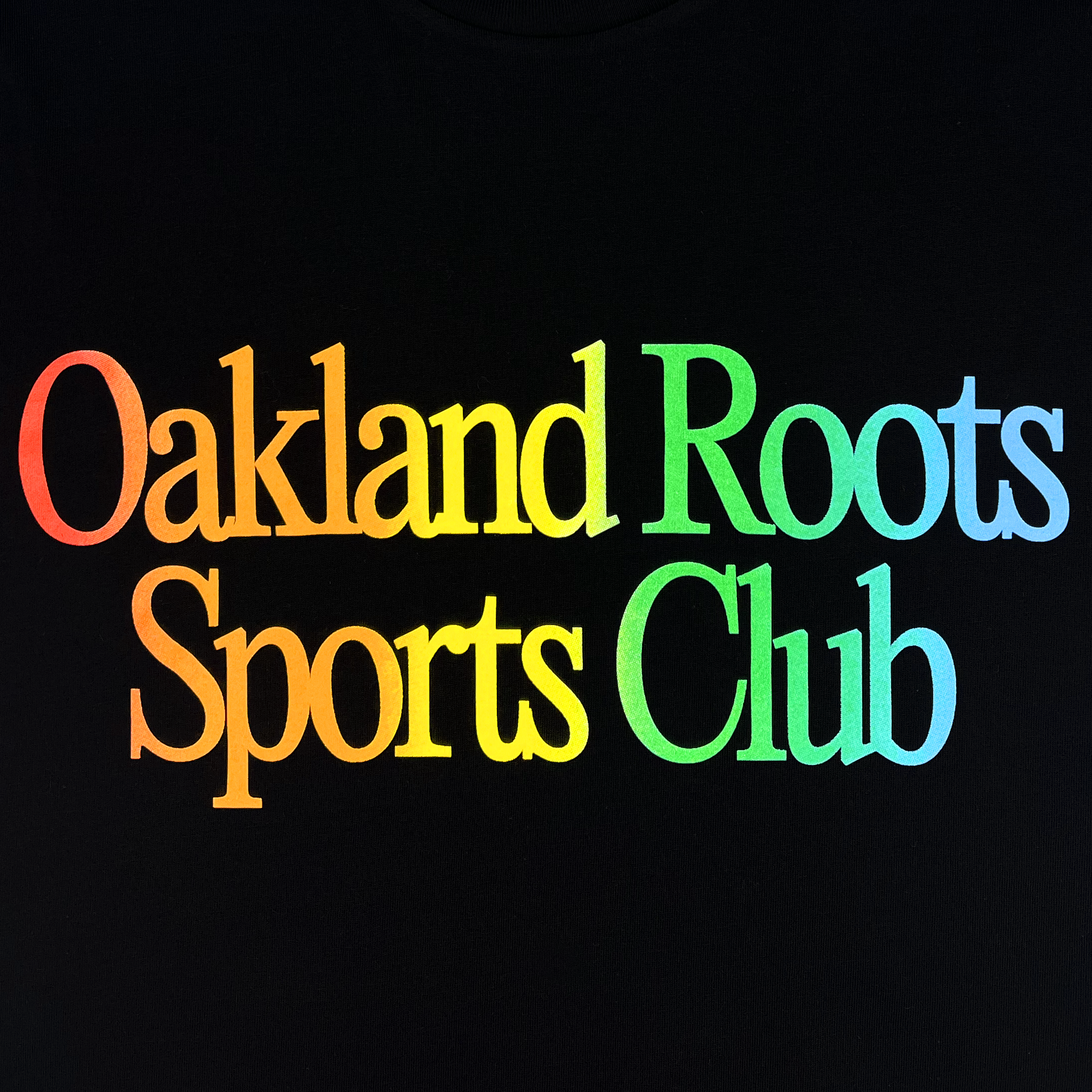 The image displays the words "Oakland Roots Sports Club" written in a bold, serif font on a black background. The text features a gradient of bright colors, transitioning from orange to yellow, green, and blue, evocative of the More Than Soccer Tee by Oakland Roots SC that embodies the spirit of social good.