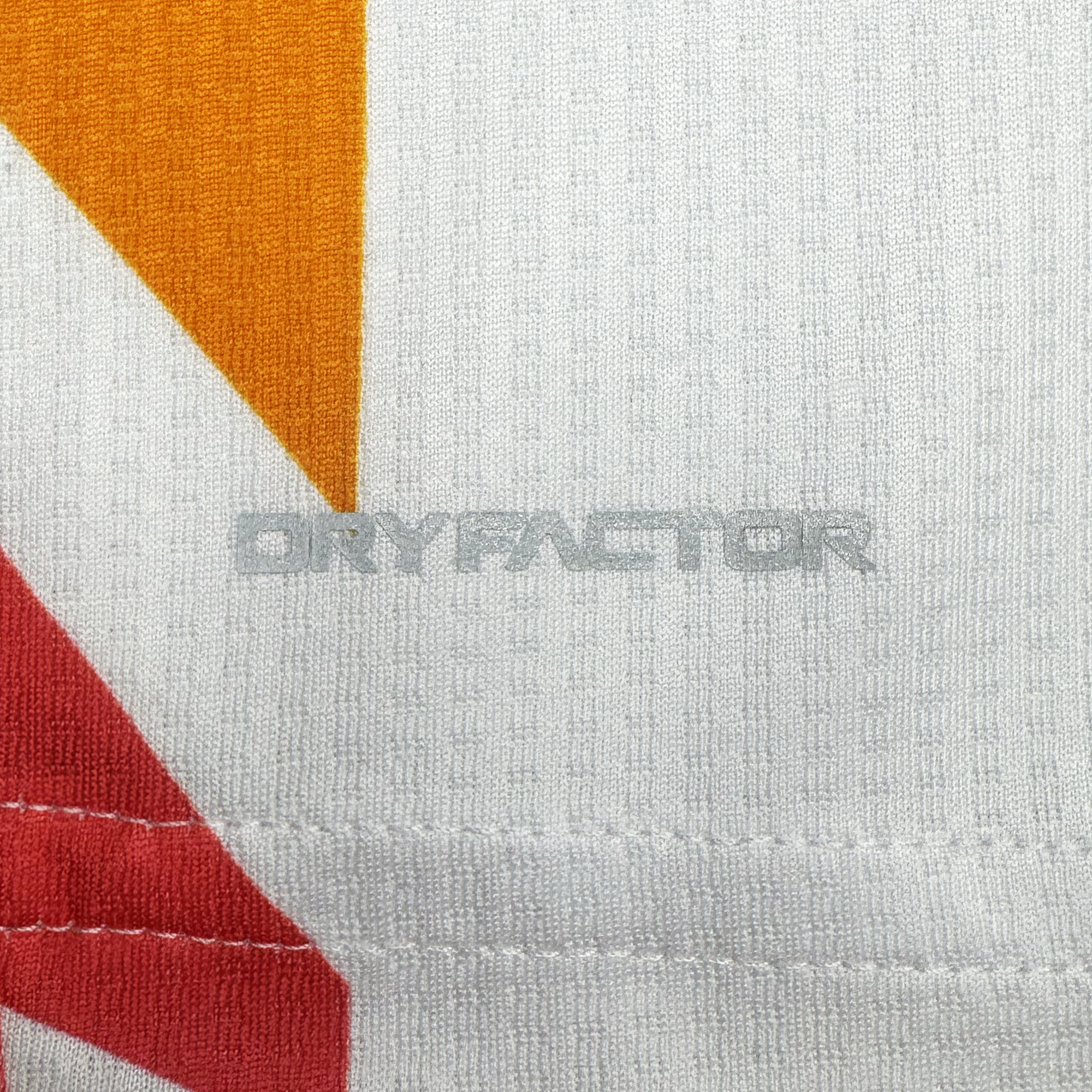 Close-up of a white fabric from the Unisex Mosaic in Motion Kit by Oakland Roots SC shows “DRYFACTOR” text, textured stitching, and bold orange-red geometric accents that highlight its mosaic-inspired soccer jersey design.