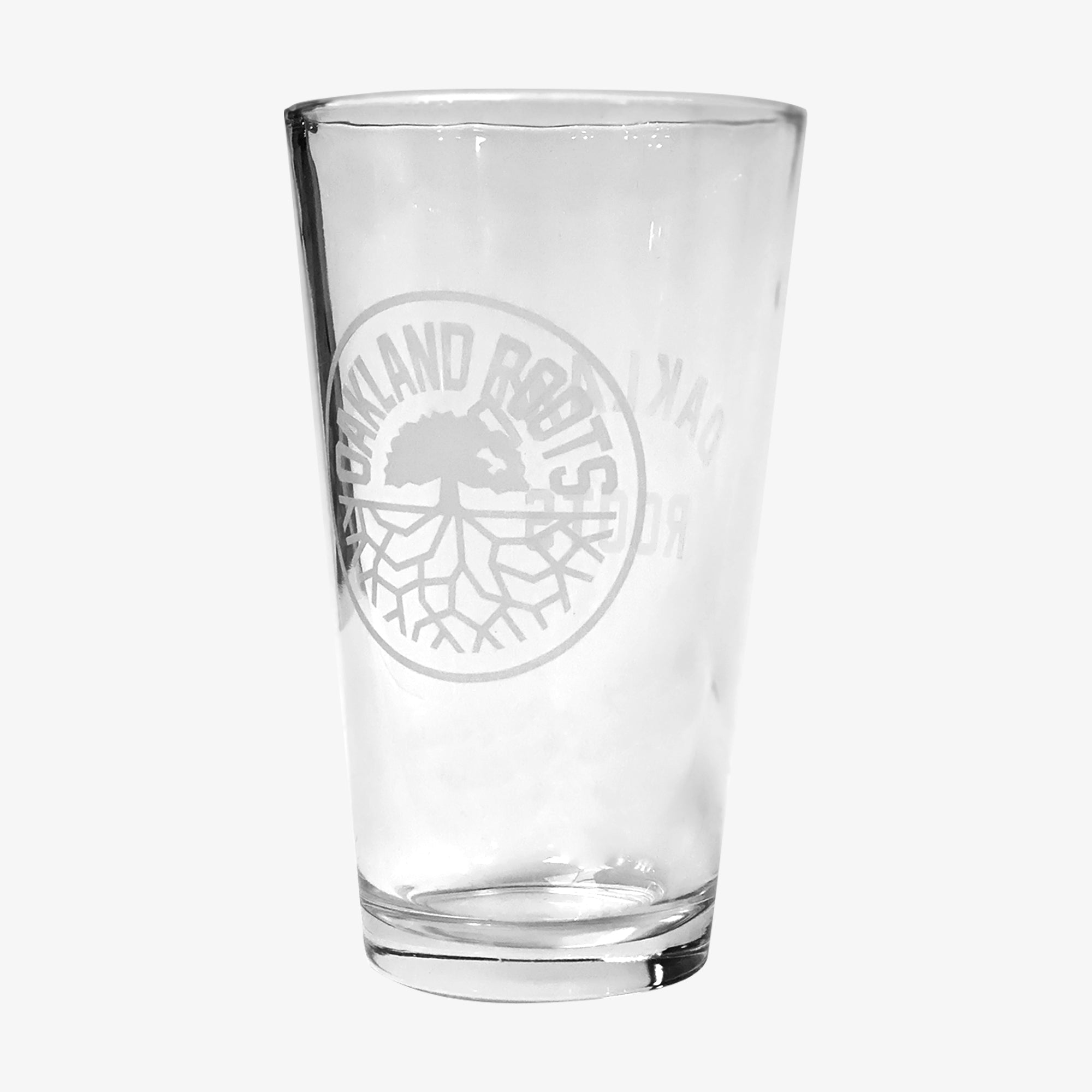 An empty Oakland Roots SC Pint Glass, branded by Oakland Roots SC, displayed against a plain white background. The clear glass features an intricate etched design of a tree with spreading roots, encircled by the words "OAKLAND ROOTS," capturing the essence of the Oakland Roots Sports Club.